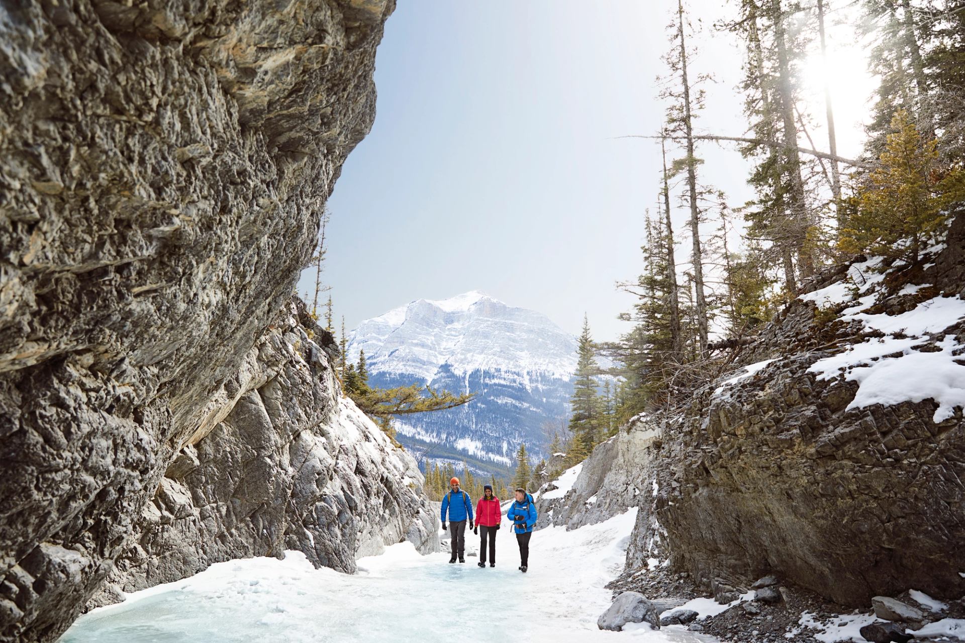 Hikers walk through a snowy canyon with rocky cliffs and distant mountain peaks..