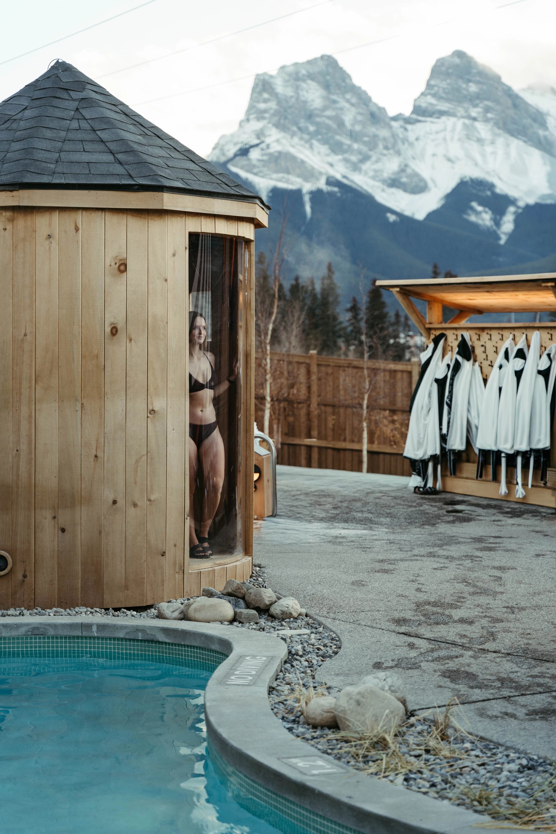 Person stepping out of a wooden sauna by a spa pool with mountain views at Everwild Canmore.