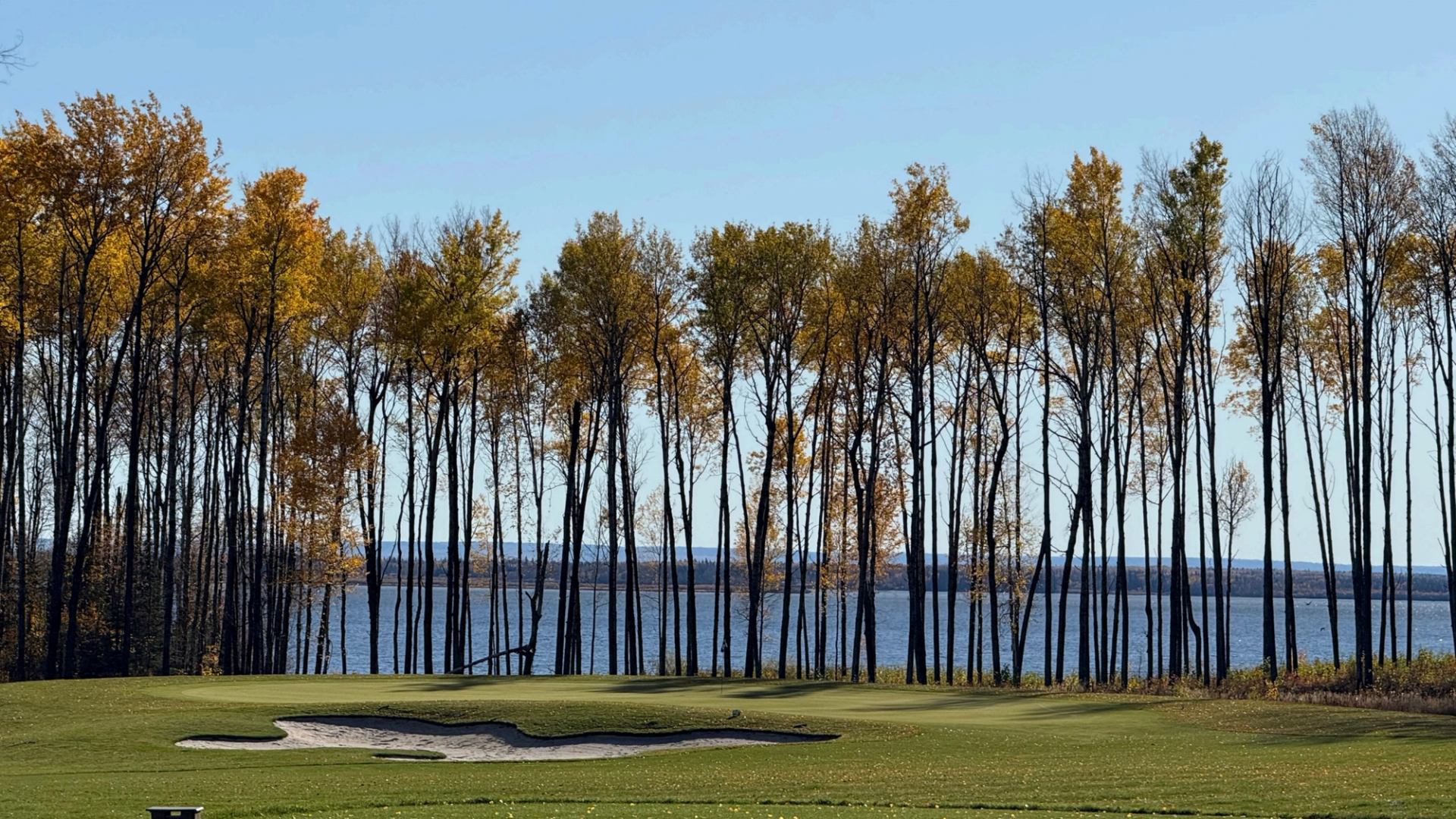 Golf course green and sand bunker beside a lake, framed by autumn trees.