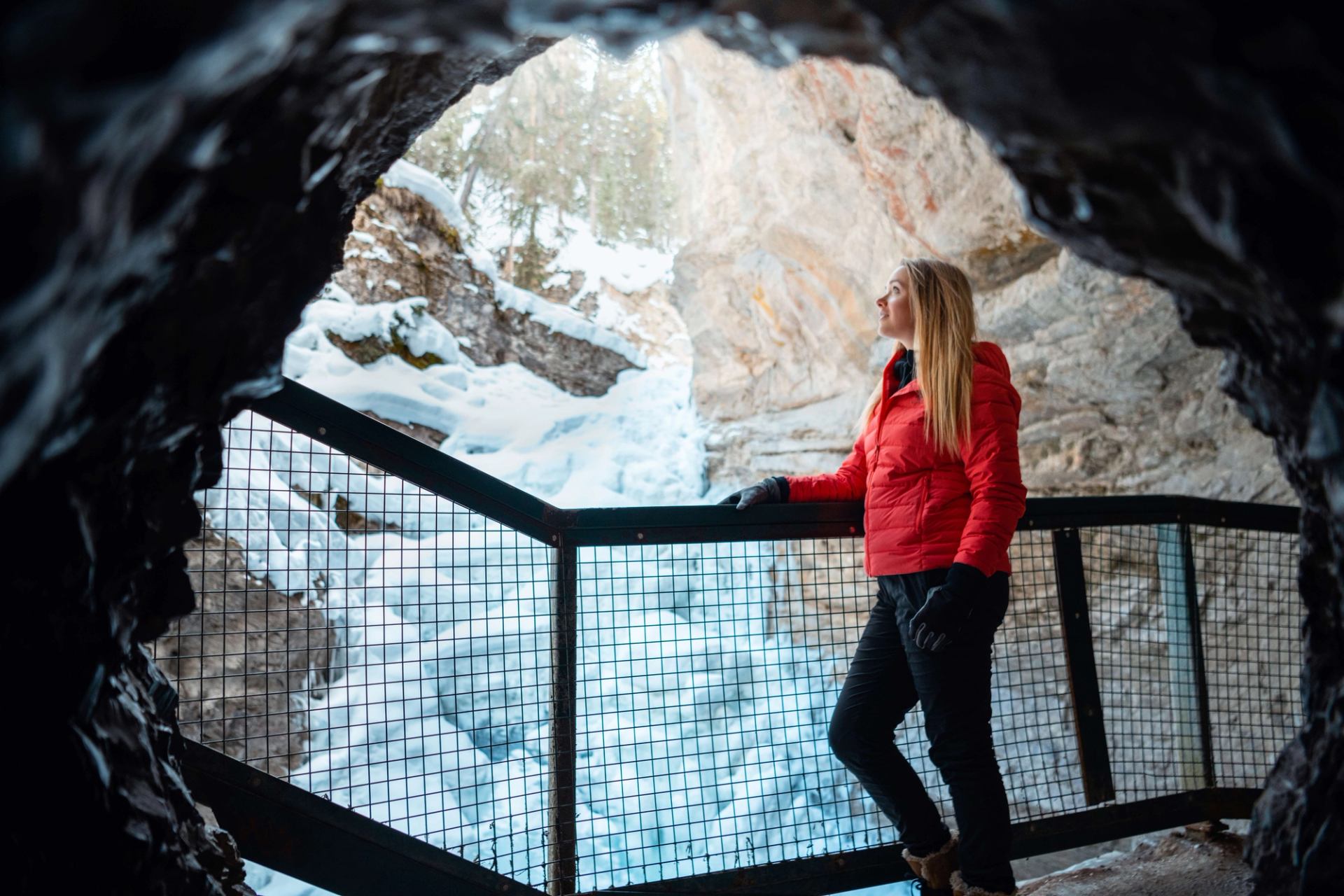 A person in a red jacket stands at a railing inside a cave overlooking a frozen canyon.