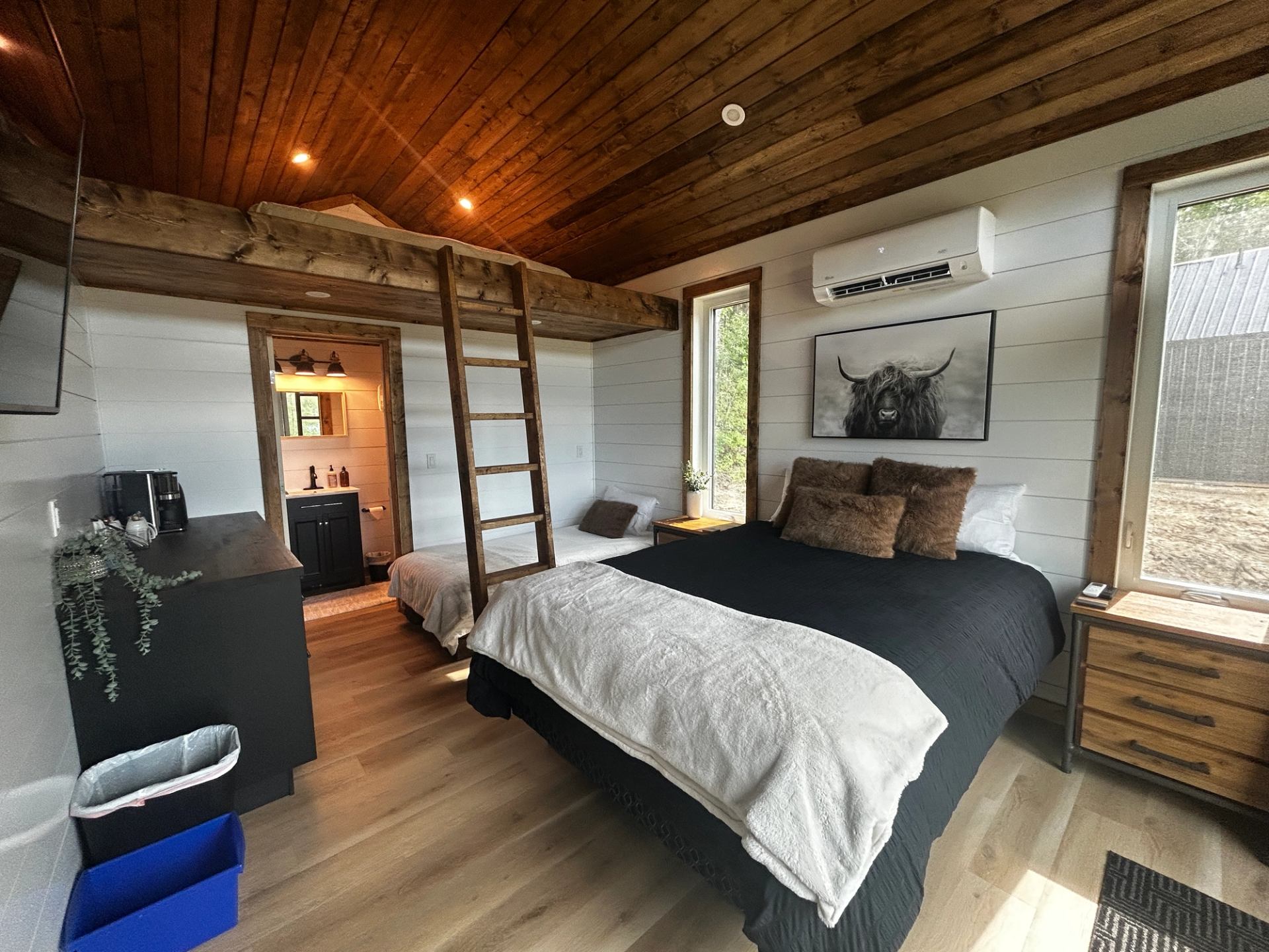 Cozy cabin bedroom with wood ceiling, large bed, loft ladder, and warm natural light from windows.