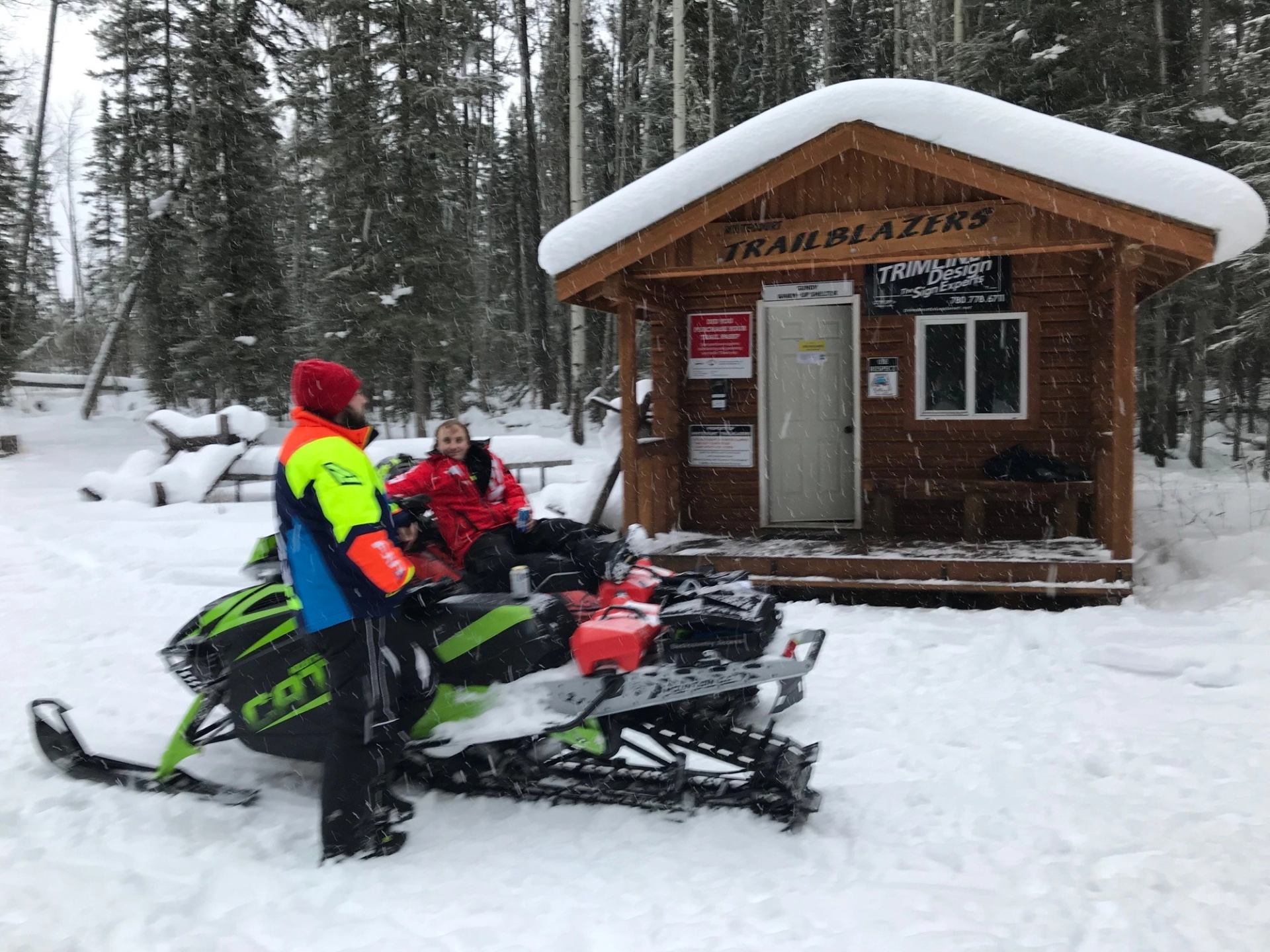 Two people in bright winter gear with snowmobiles stand by a small wooden cabin in snowy forest, evoking adventure and warmth.