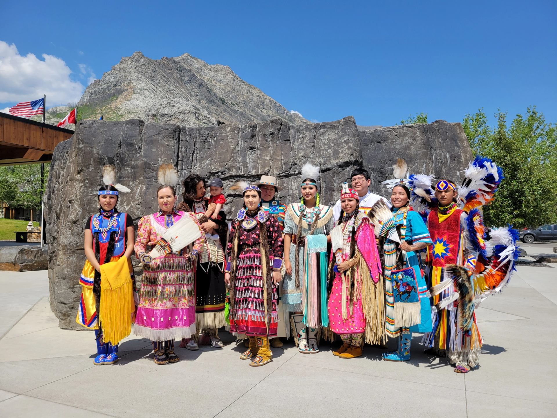 A group of Indigenous people dressed in vibrant traditional regalia pose in front of a large rock with a mountain in the background. The scene is set outdoors on a sunny day, with flags and greenery visible.