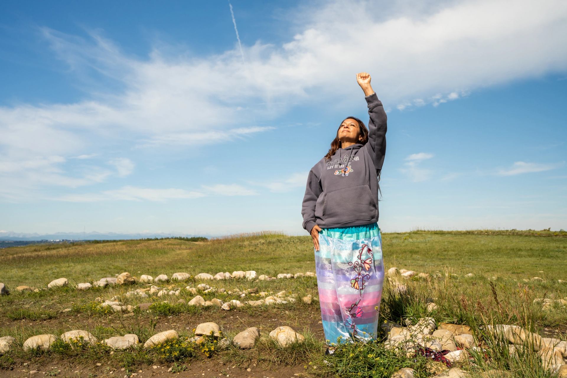 A person stands in a grassy stone circle, raising one arm under a bright blue sky.