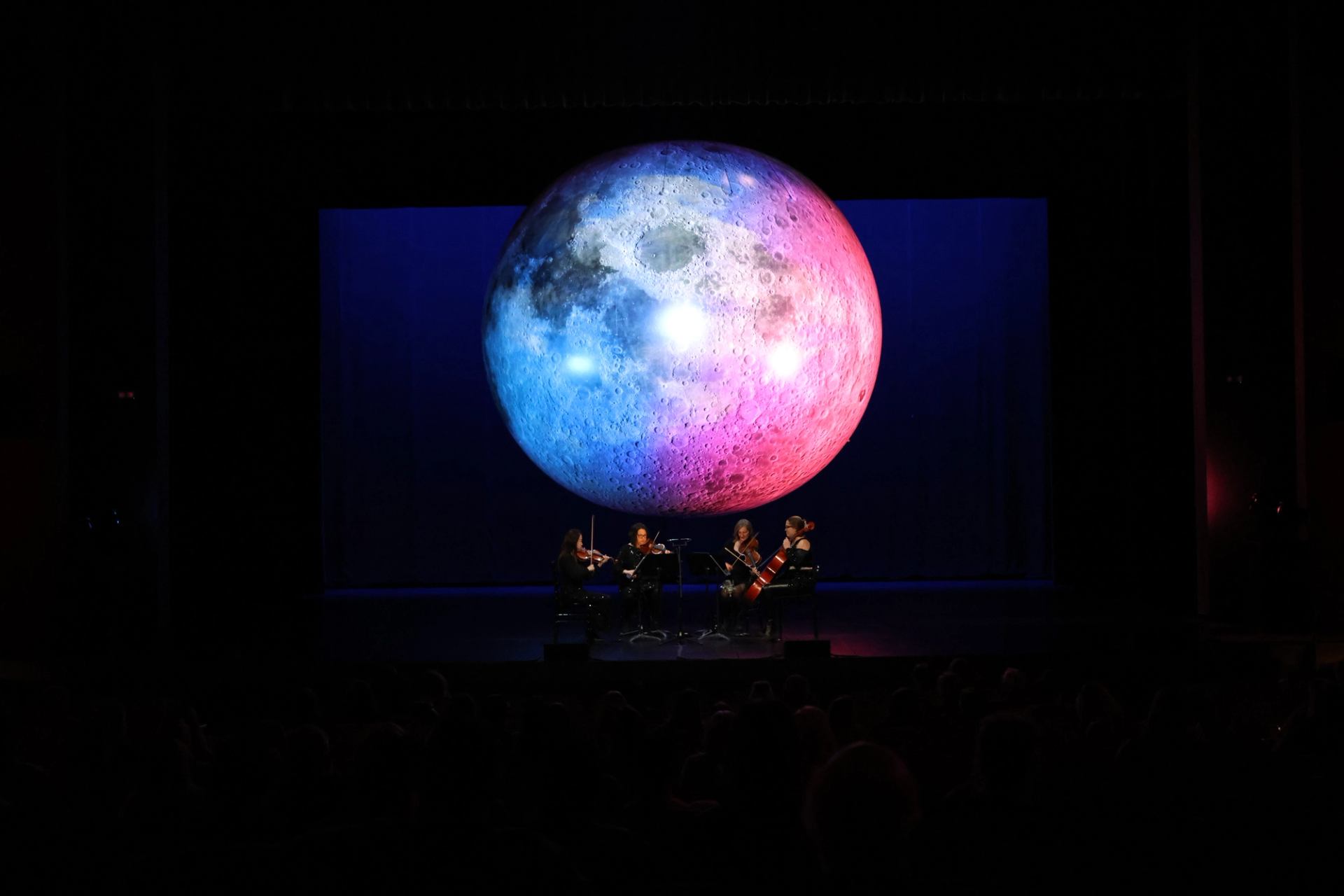 String quartet performs on dark stage beneath large blue and pink moon projection.