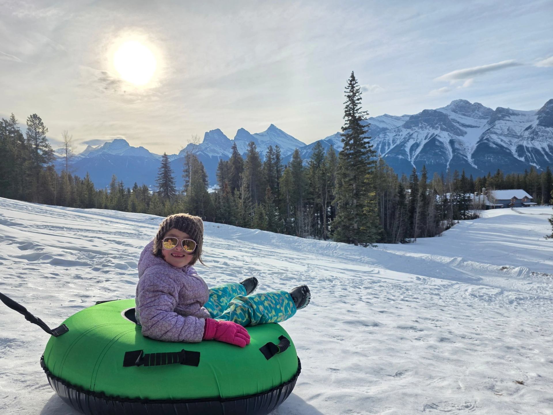 Person sitting on a green snow tube on a snowy slope with pine trees and mountain peaks at Silvertip Resort.
