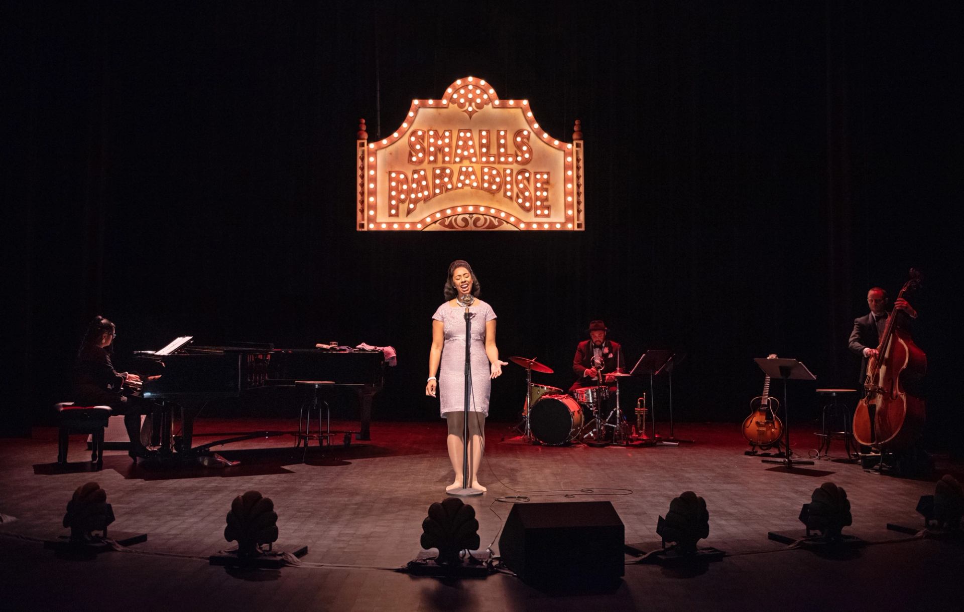 Jazz singer performs onstage with pianist, drummer and bassist under Smalls Paradise marquee.