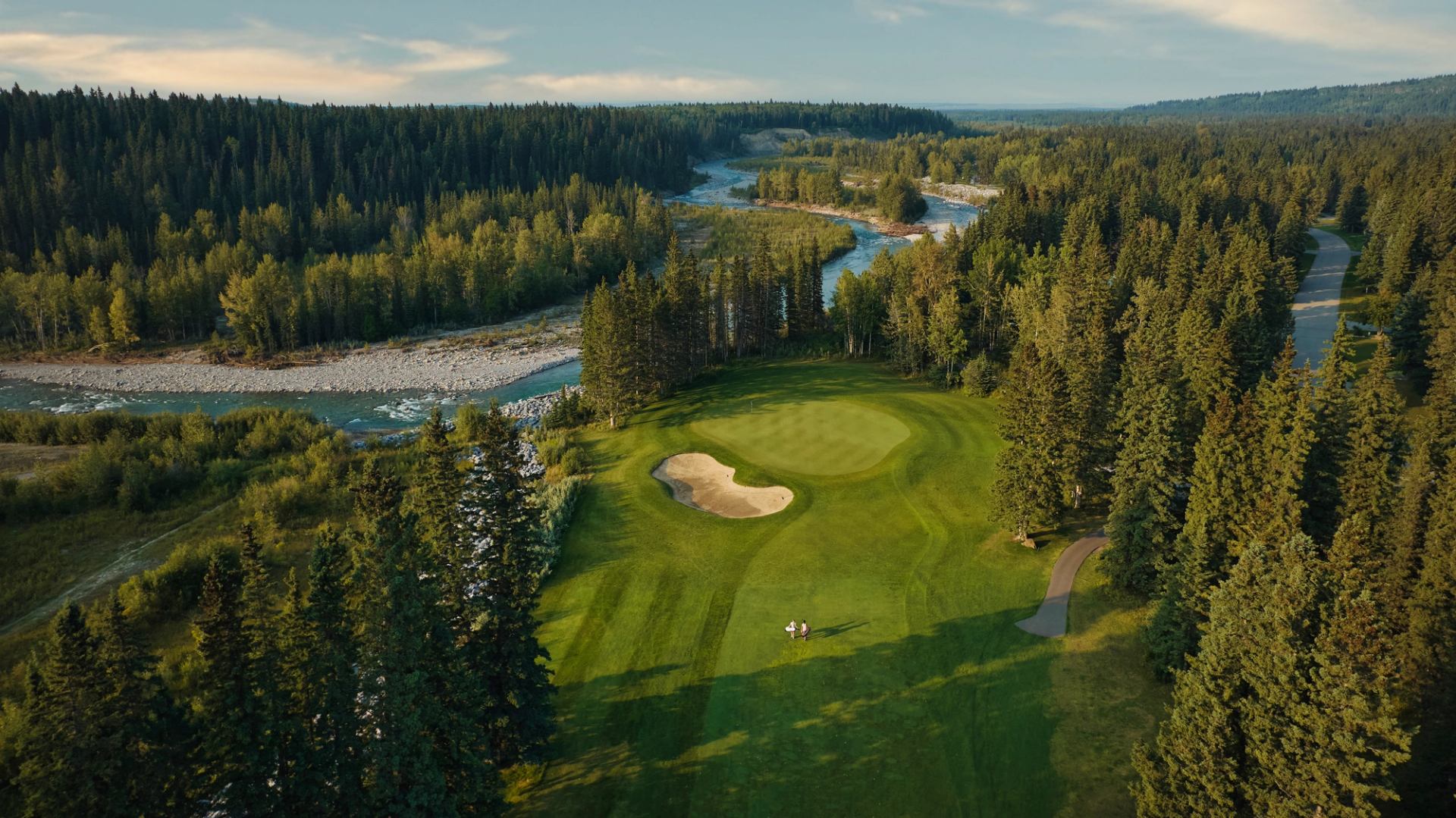 Golf green with sand bunker beside a winding river and dense evergreen forest.