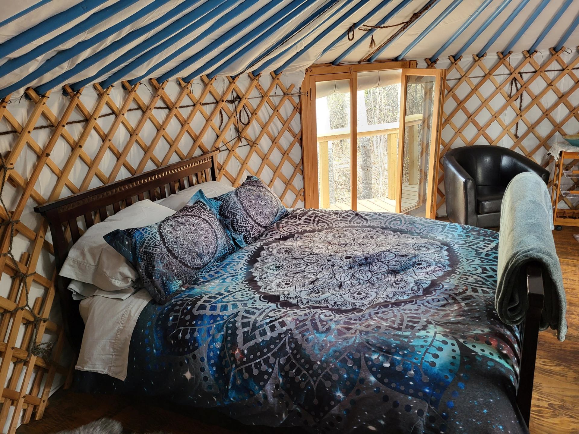 Interior of a yurt with a bed featuring a blue mandala-galaxy duvet, wooden lattice walls, and a large window.