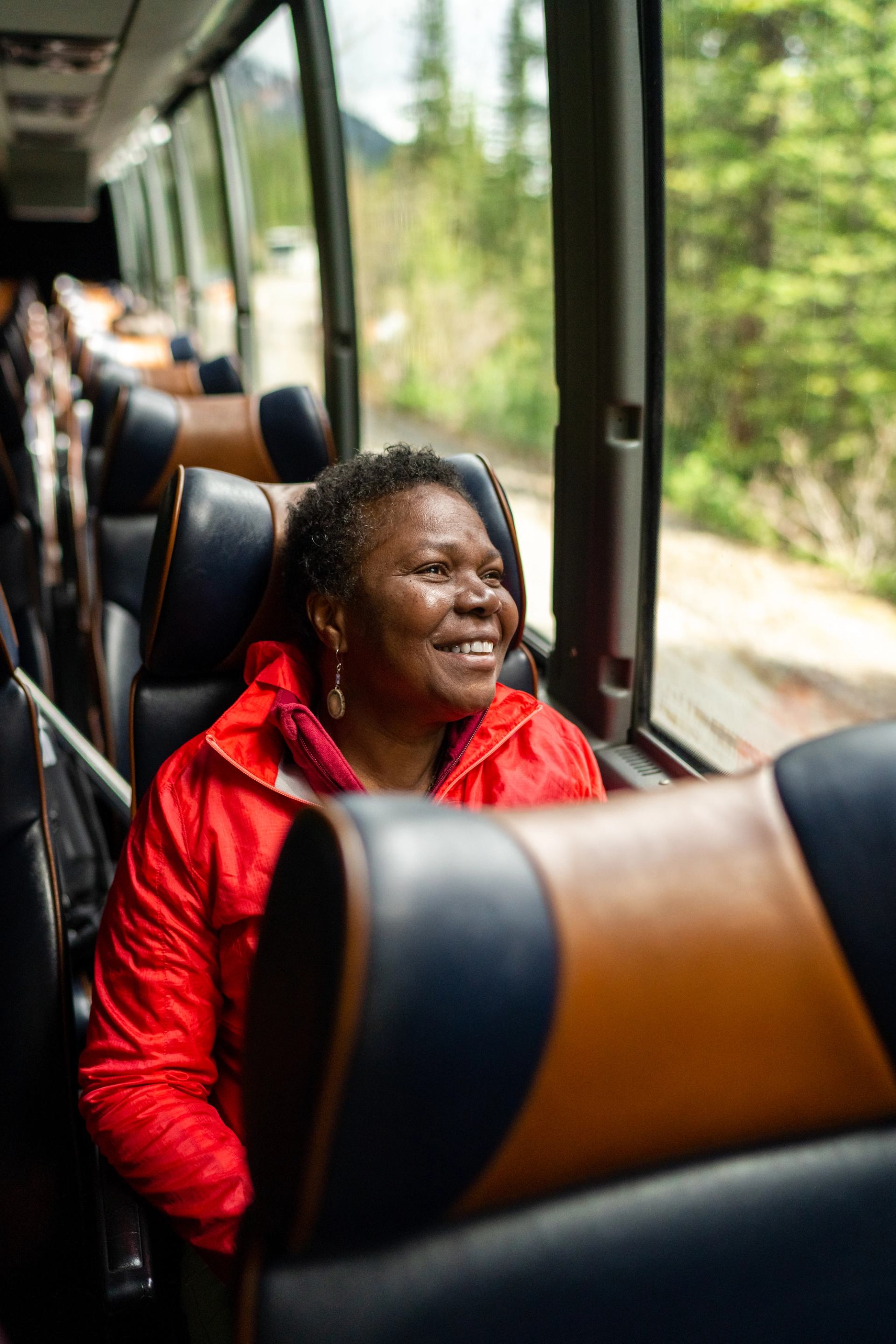 Passenger seated on a shuttle bus wearing a bright red jacket, looking out a large window at a forested mountain landscape with trees and a dirt road visible outside; rows of leather seats line the bus interior.