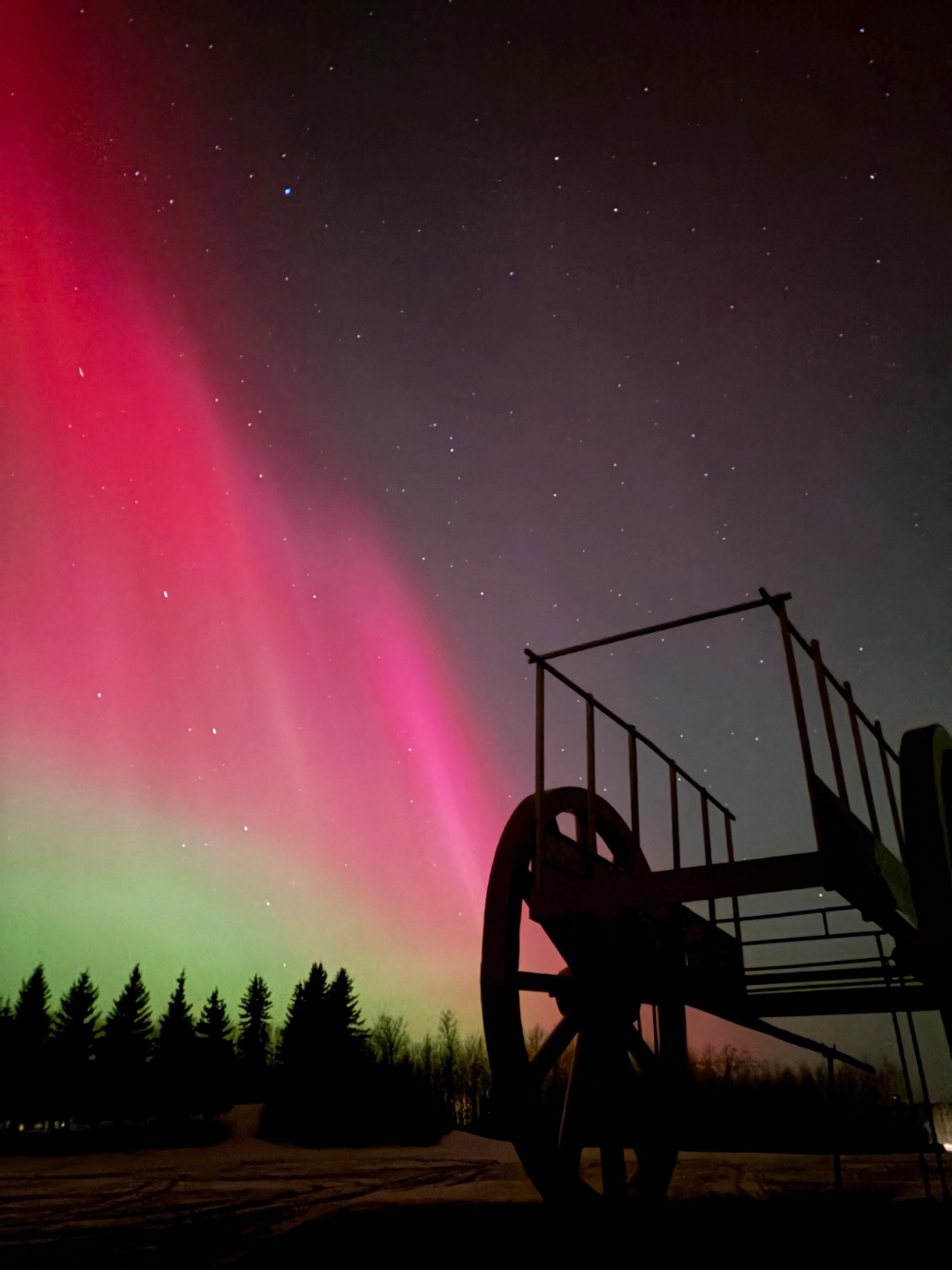 Silhouette of Alberta’s Largest Red River Cart under vivid red and green northern lights.