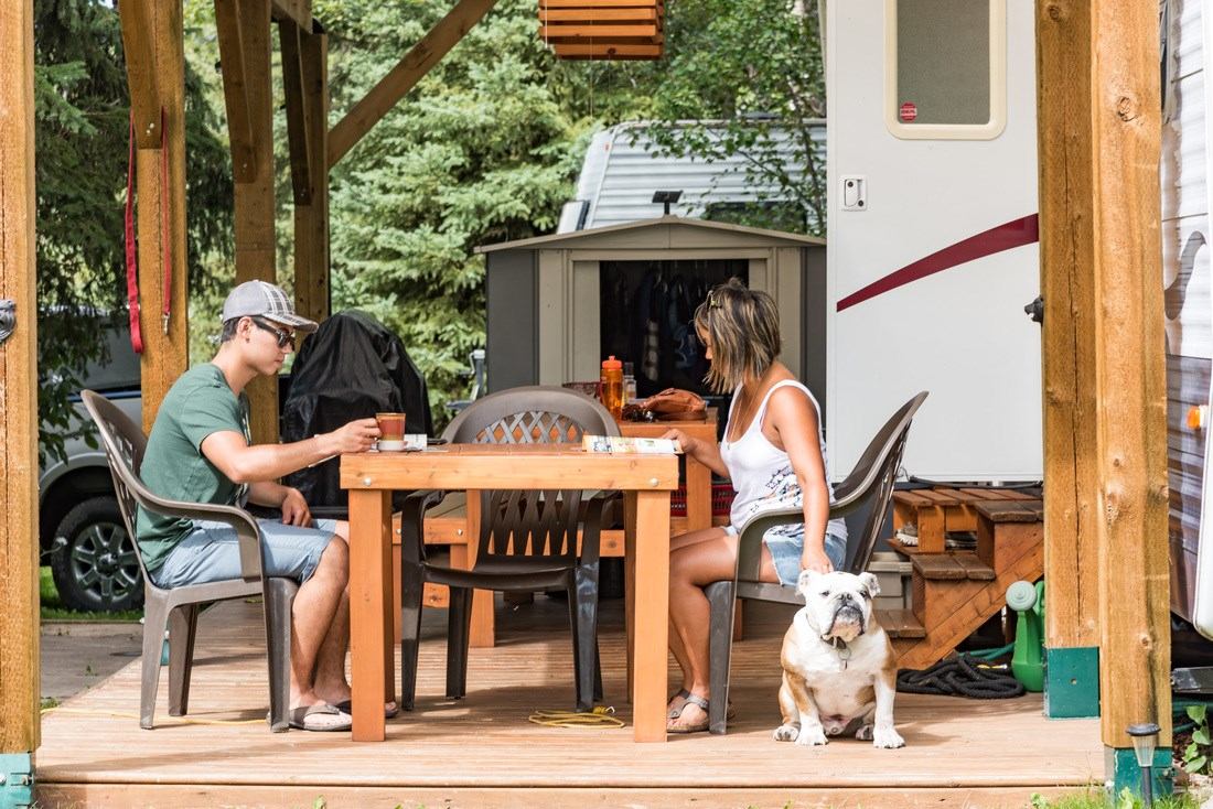 Two people sit at a wooden patio table near an RV, with a bulldog beside them.