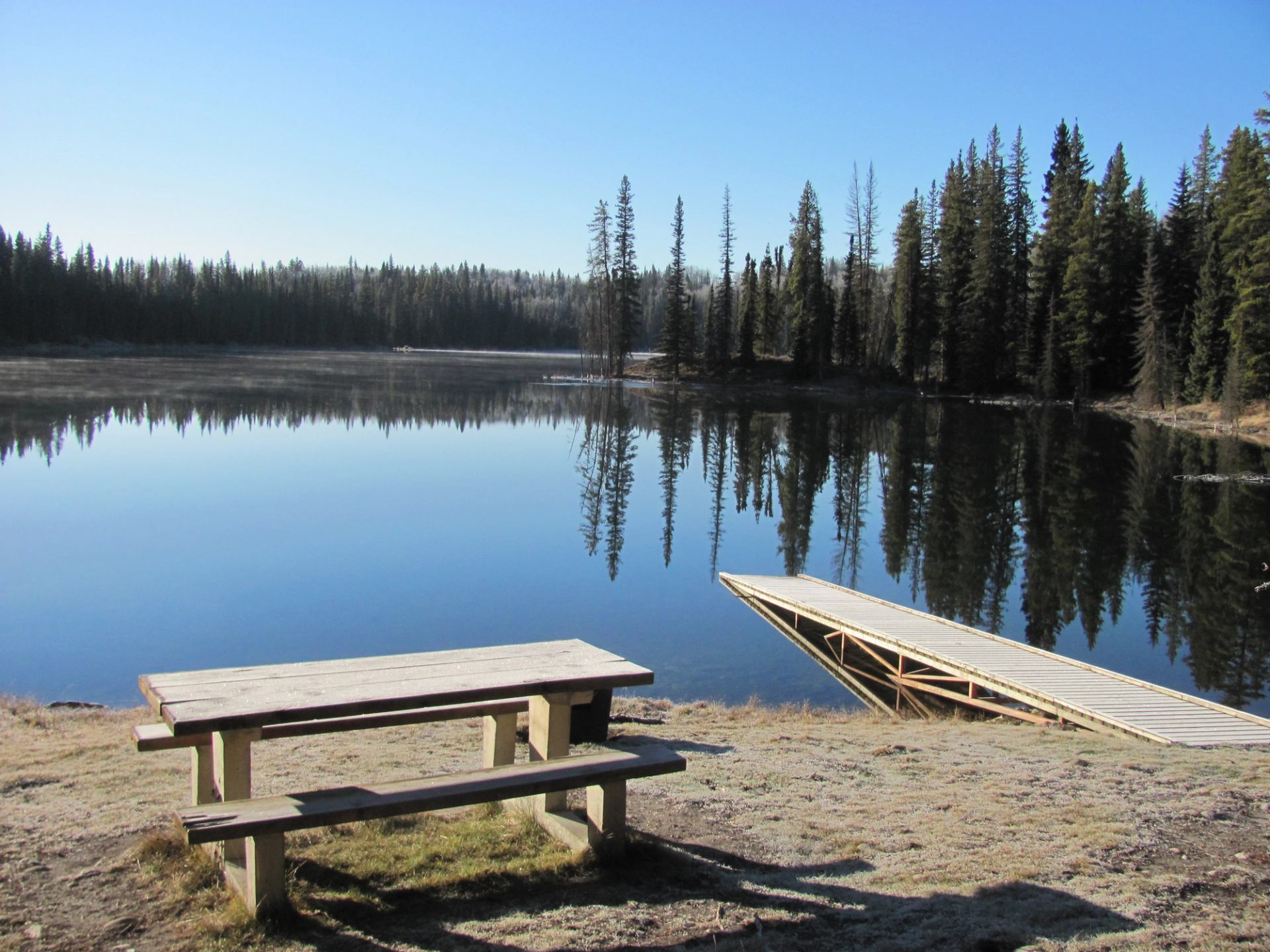 Wooden picnic table and dock beside calm forest lake reflecting tall evergreen trees.