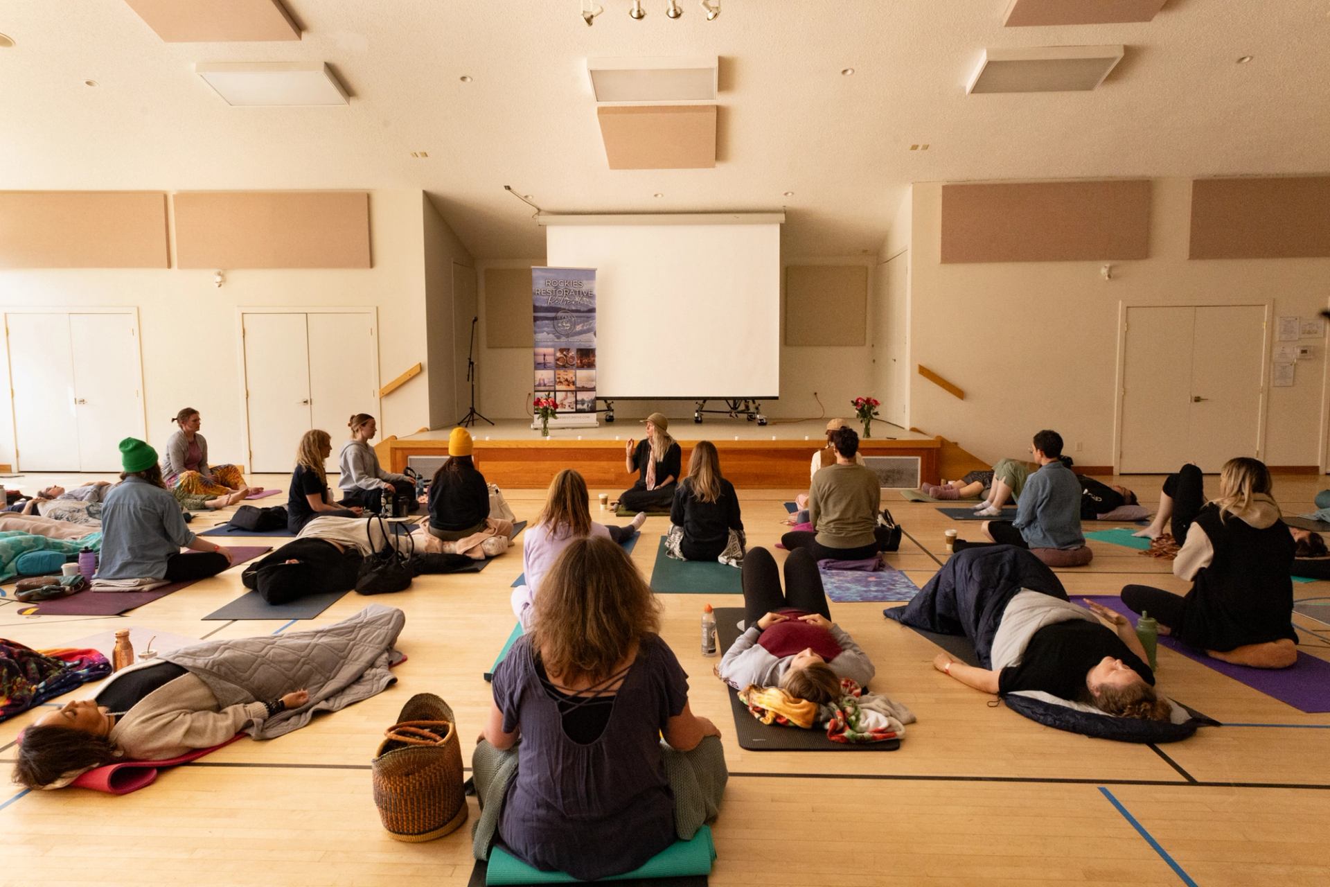 People on yoga mats sit or lie in a community hall facing a projector screen.