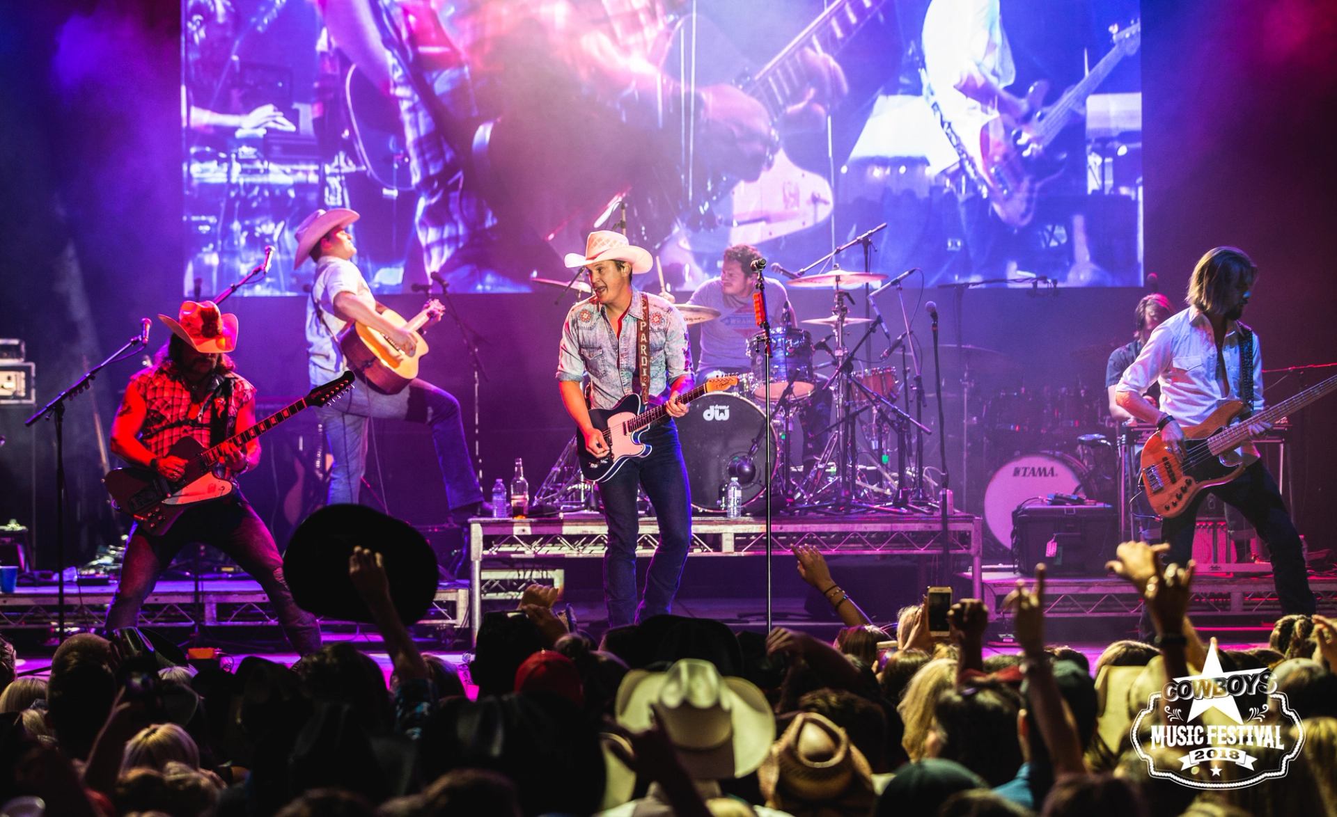 Country band performing on a brightly lit stage with crowd in front at Cowboys Music Festival.
