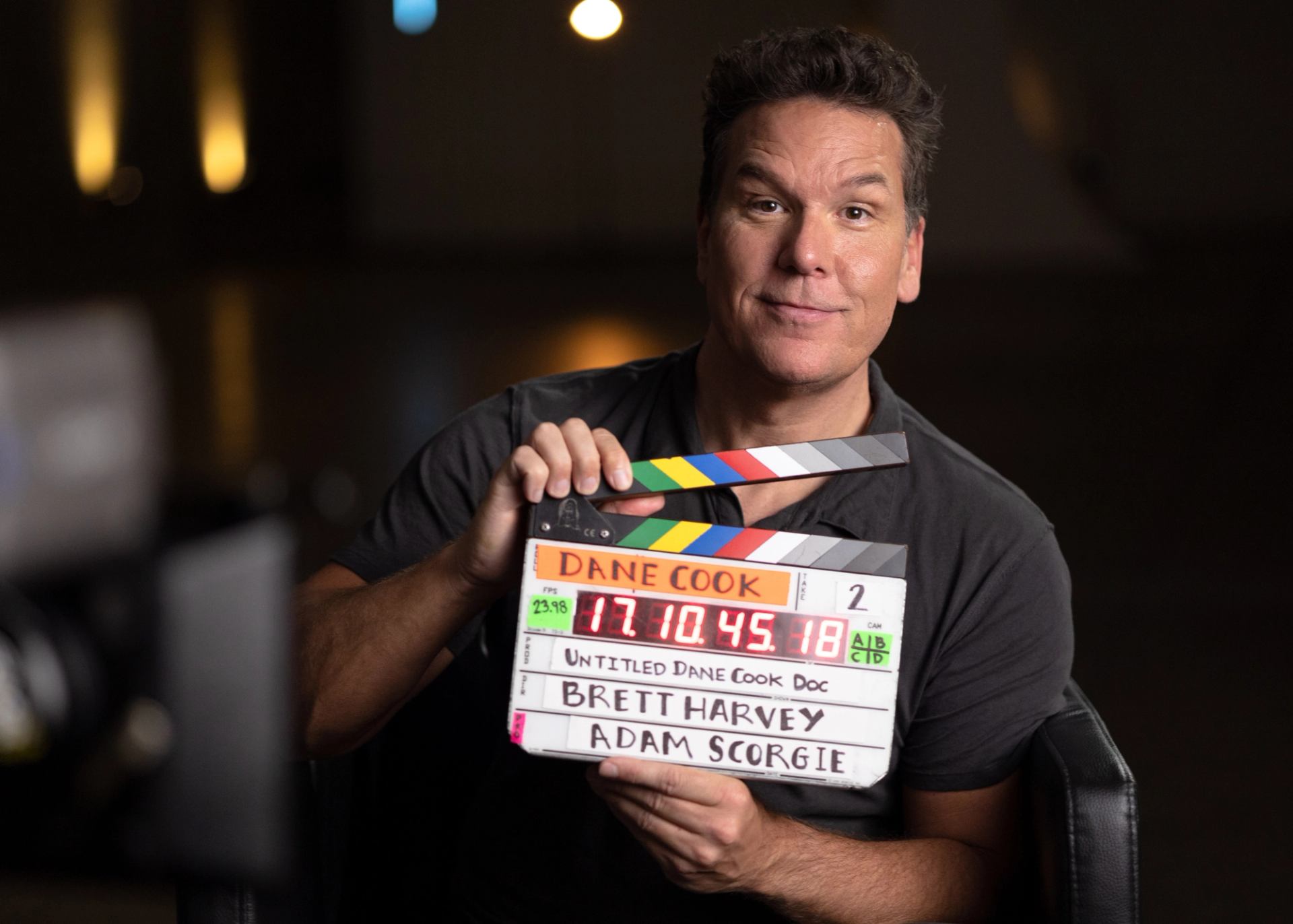 Person holding colourful film clapperboard labelled Dane Cook in dim studio setting.