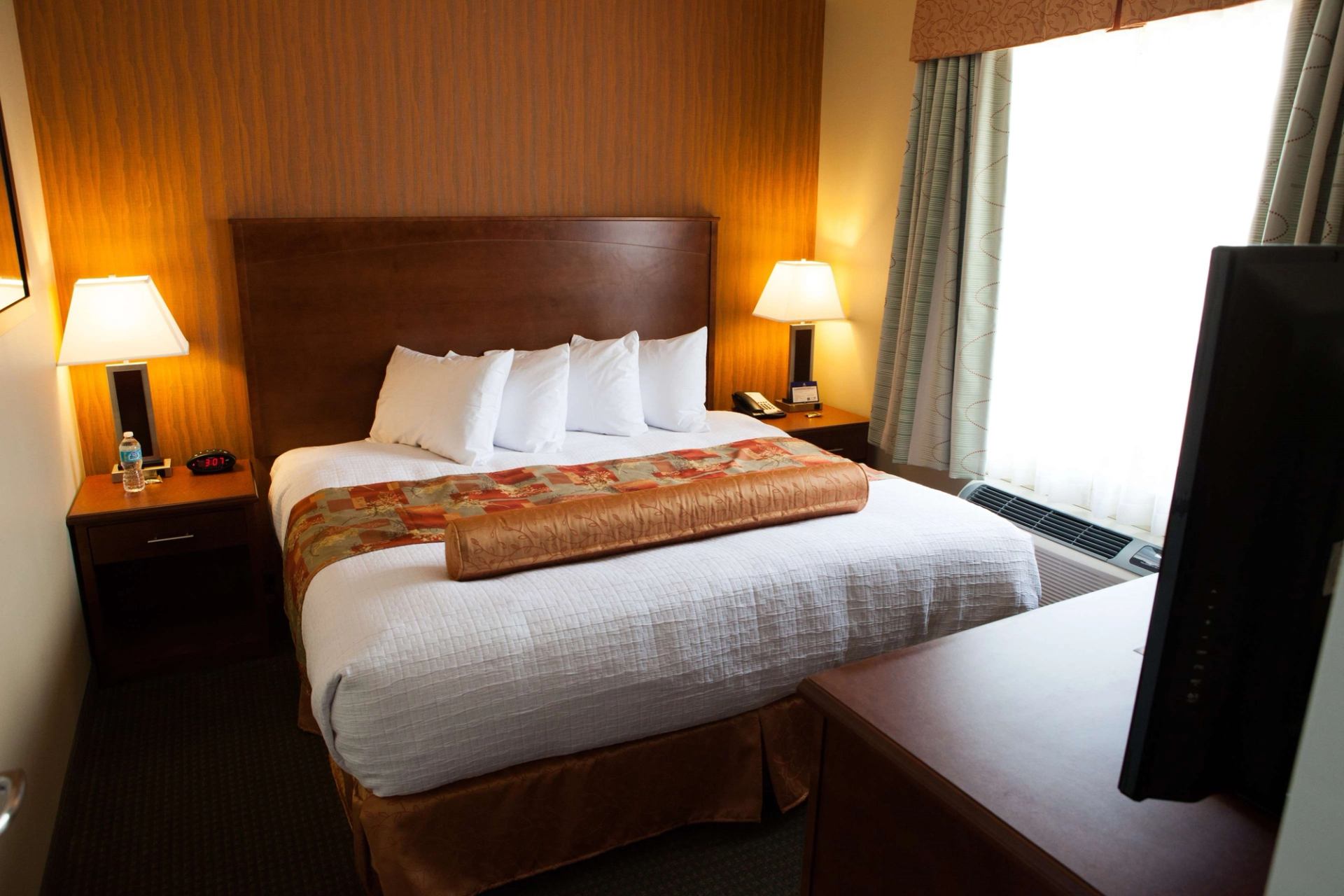 Hotel room with king bed, bedside lamps, TV, and window with curtains.