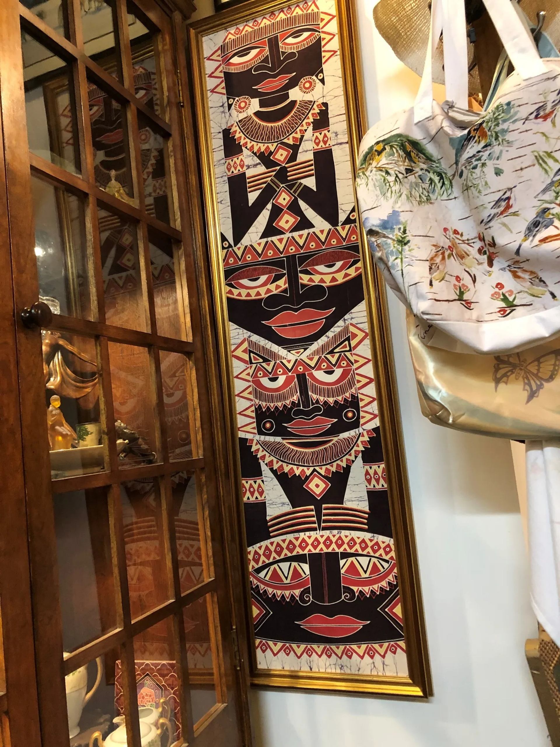 Tall framed artwork with stylized geometric faces beside glass cabinet and patterned tote bag.