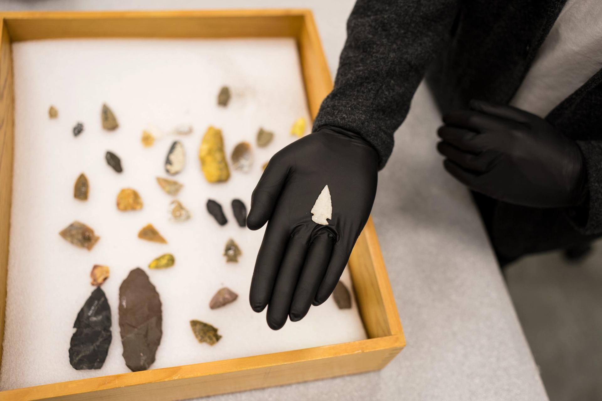 Gloved hand holding small white arrowhead above tray of assorted stone artefacts.