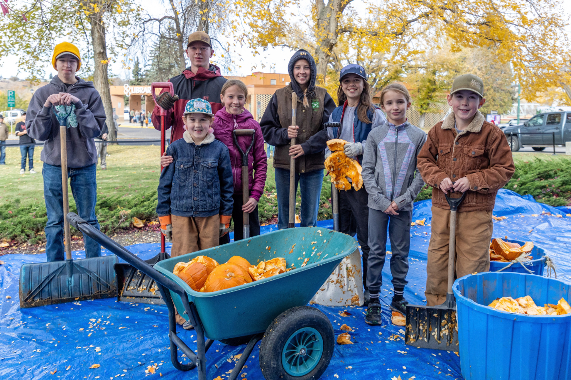 Group holding shovels beside a wheelbarrow and bins filled with smashed pumpkins.