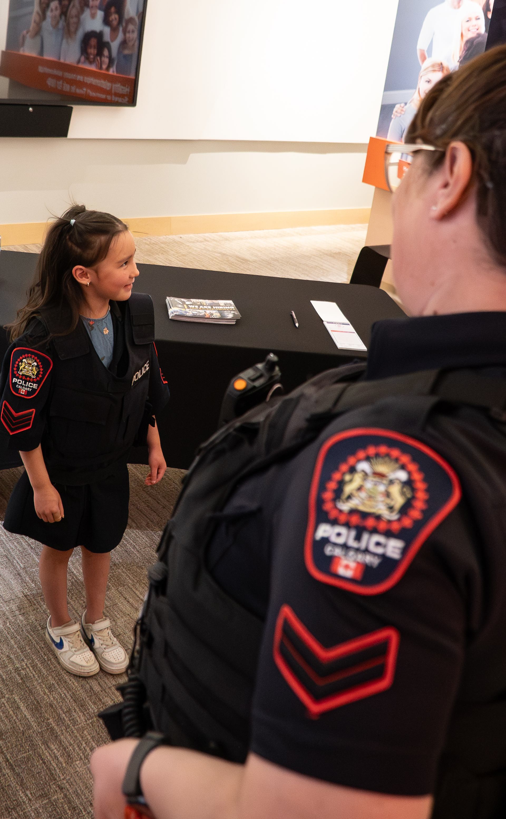 Child wearing Calgary police vest stands near officer at indoor information table.