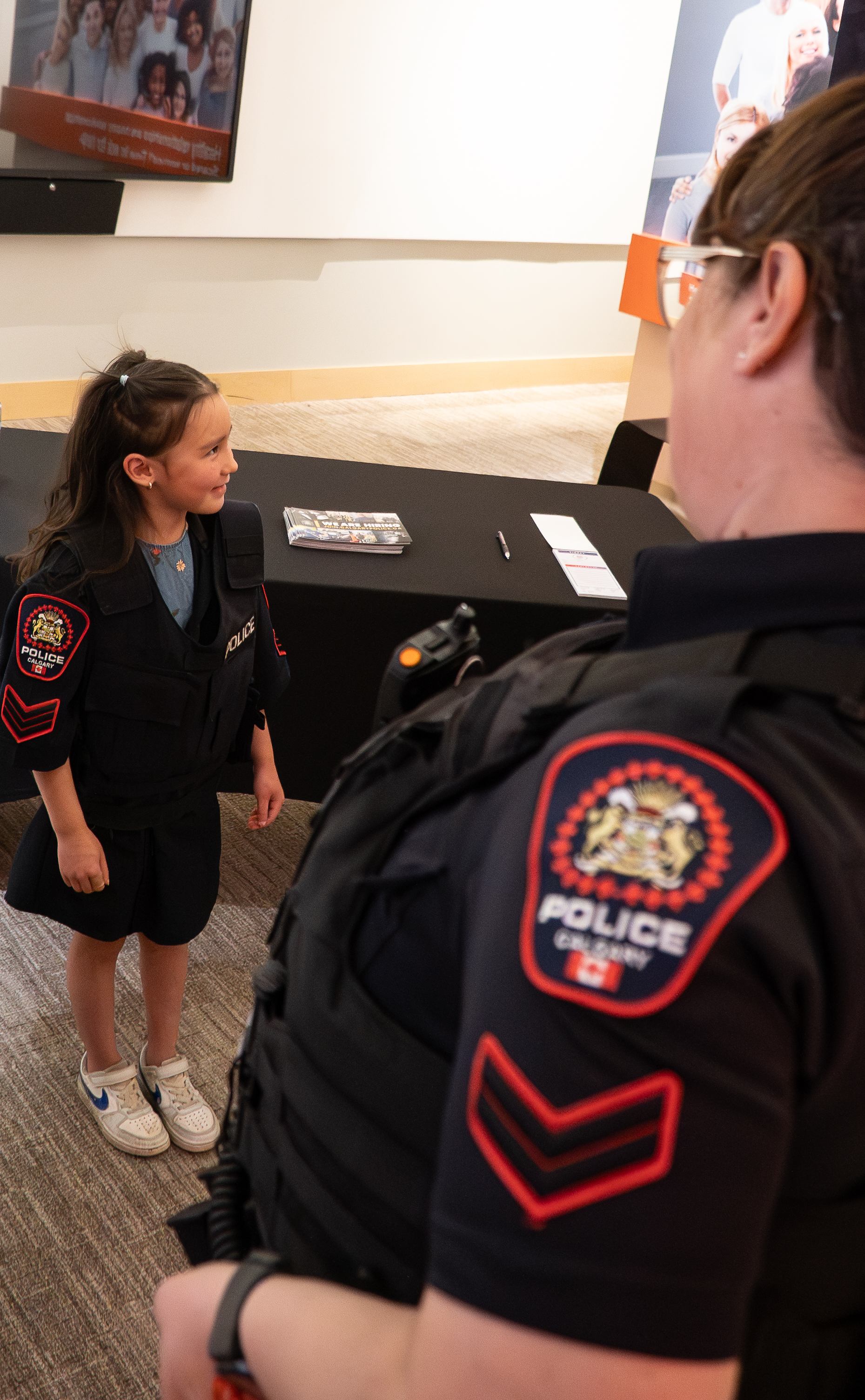 Child wearing Calgary police vest stands near officer at indoor information table.
