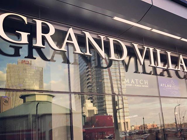 Grand Villa sign on glass facade with city buildings reflected.