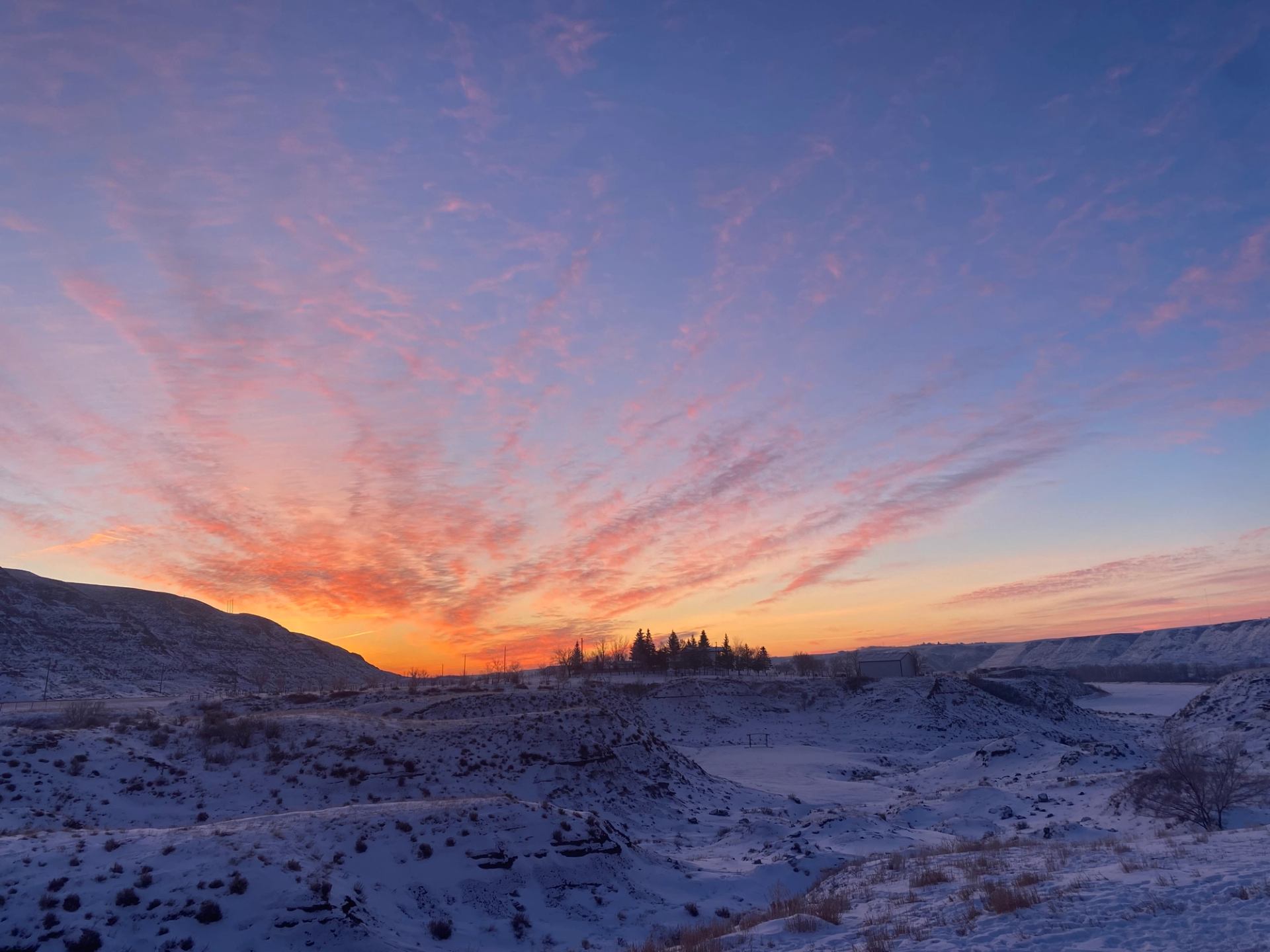 Vibrant winter sunset over snowy hills with trees silhouetted on the horizon.