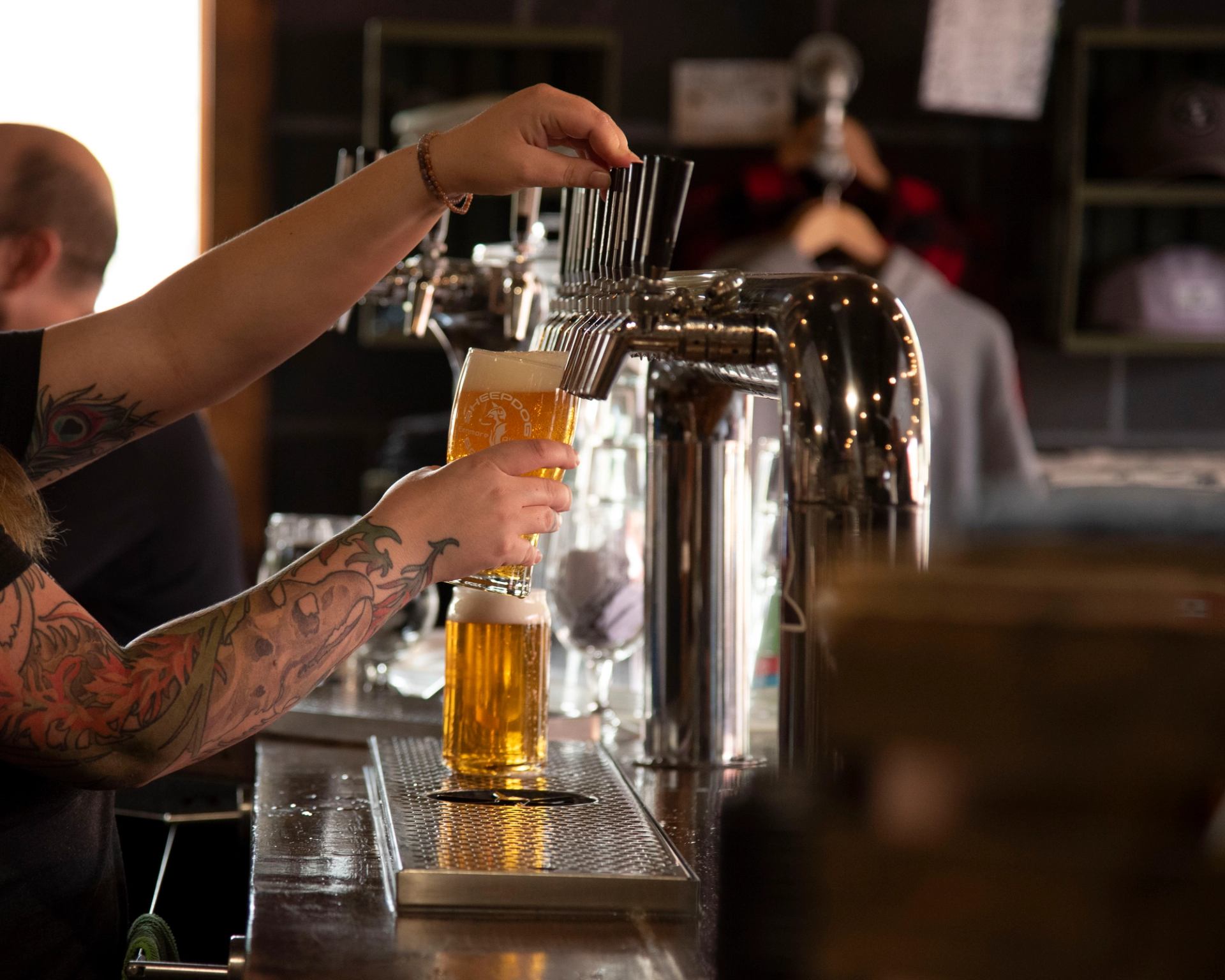 Tattooed bartender pours golden beer from a tap into a tilted glass.