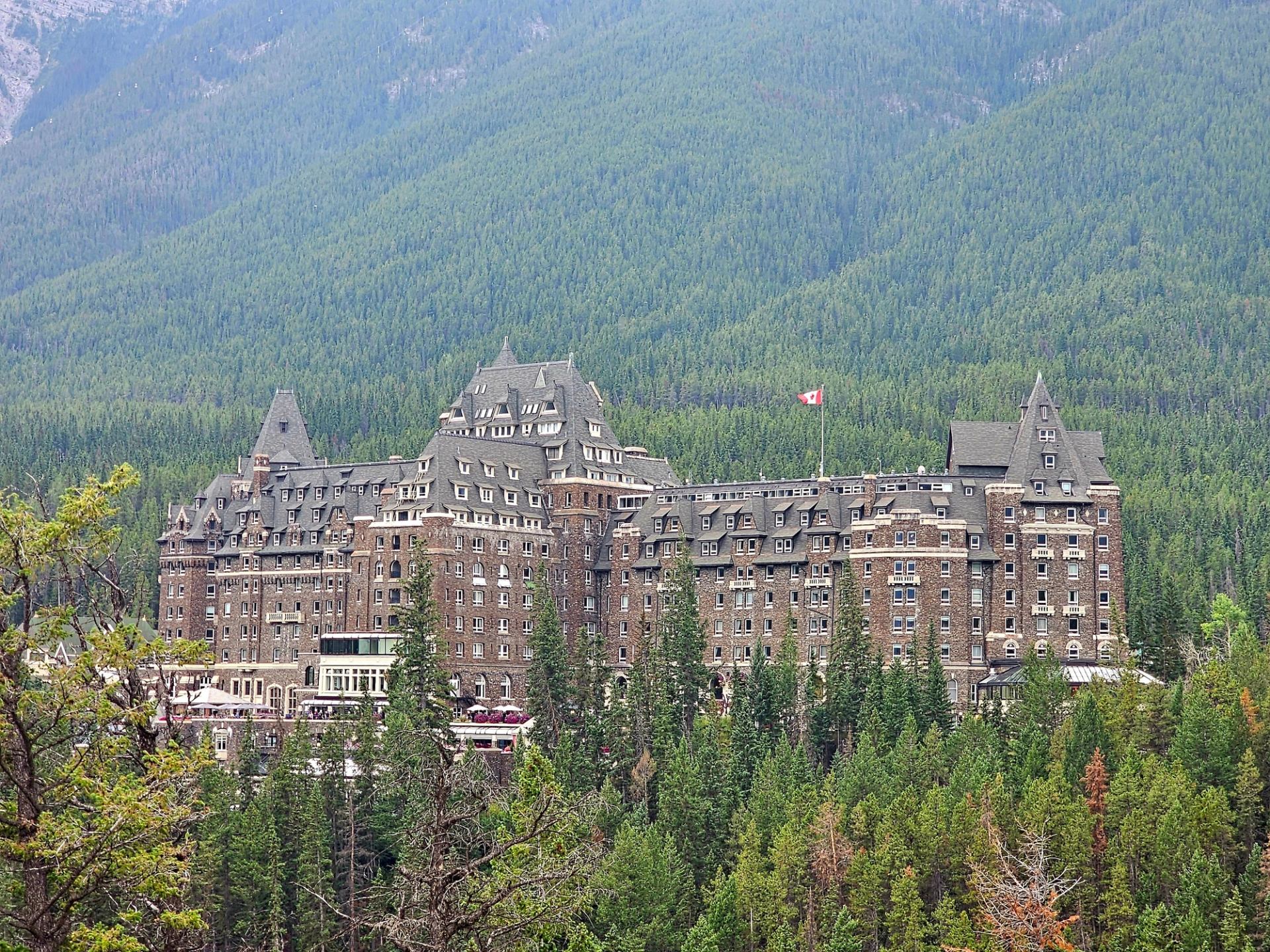 Large stone hotel with Canadian flag surrounded by dense evergreen forest and mountains.