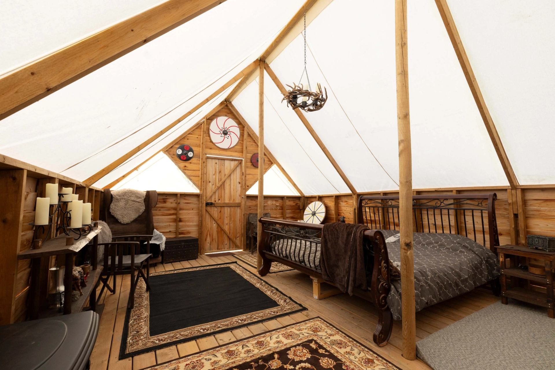 Interior of a spacious glamping tent with a dark wooden bed, armchair, and rustic decor including decorative shields on the back wall.