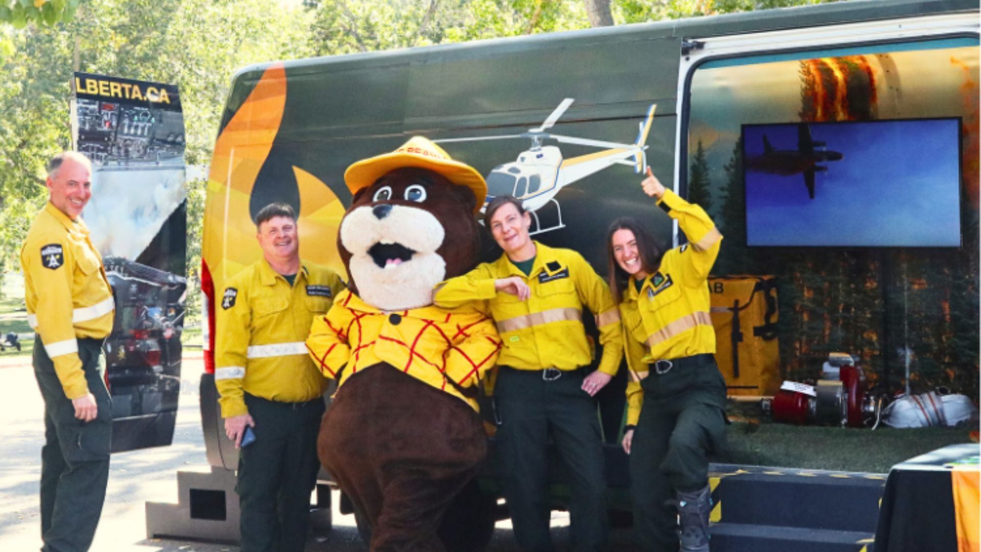 A group of Alberta Wildfire team members in yellow uniforms stand smiling with Bernie the Beaver, the official wildfire safety mascot, in front of a display van at the Alberta Biodiversity Festival.