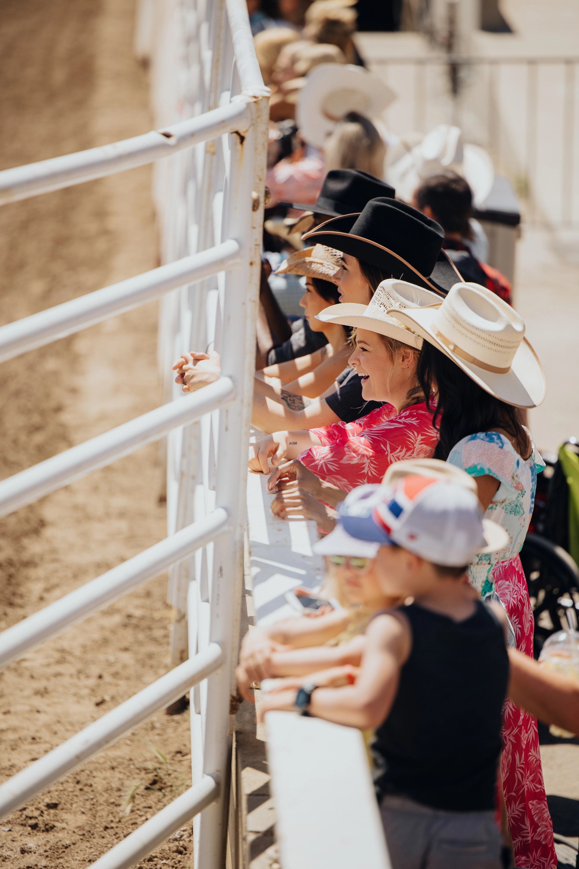 Spectators in cowboy hats lean on a white fence overlooking a dirt arena.