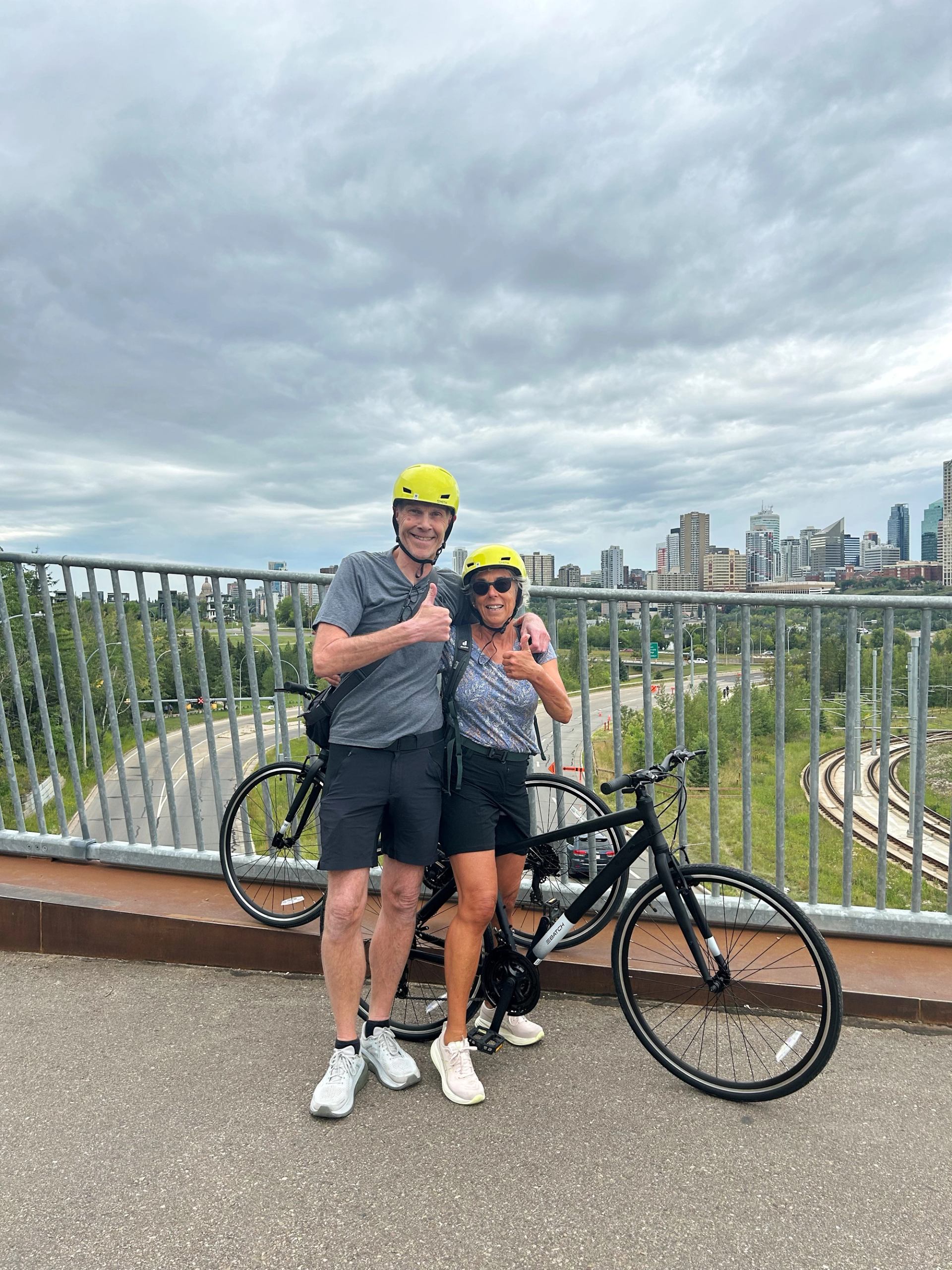 Two helmeted cyclists stand on bridge with bike, city skyline in background.