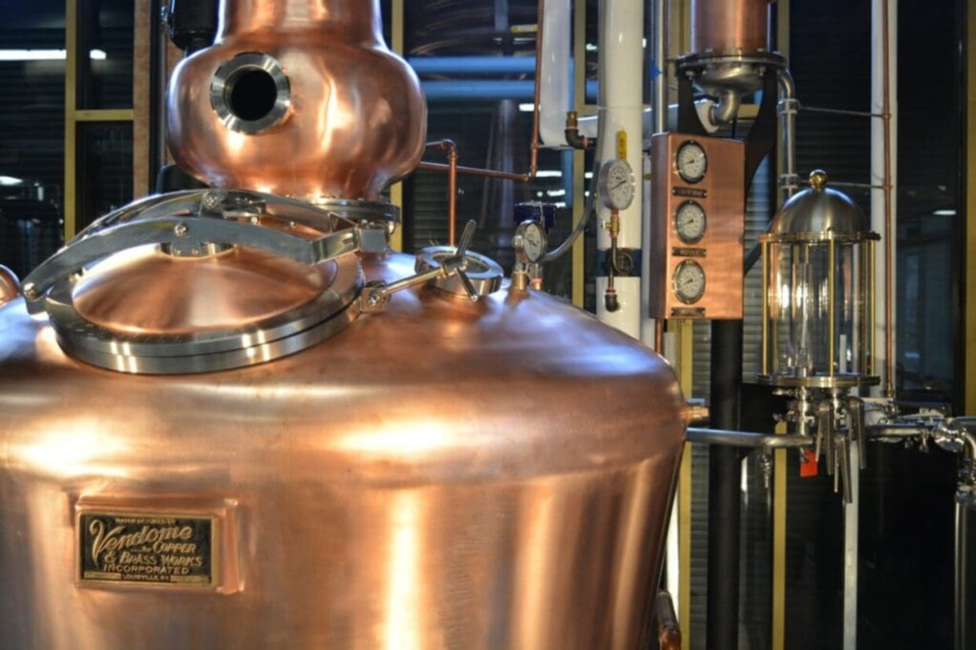Copper distillation tank with gauges, pipes and glass column inside an industrial facility.