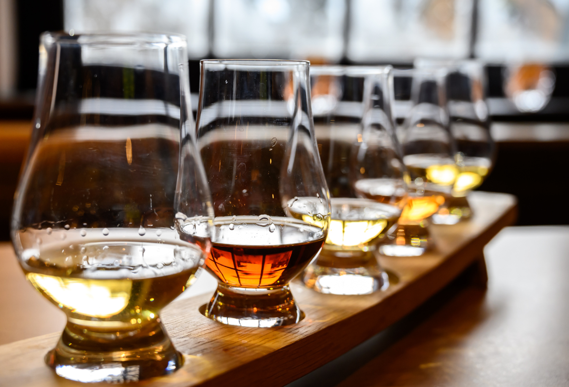 Whisky tasting glasses with amber and golden samples line a wooden flight board.