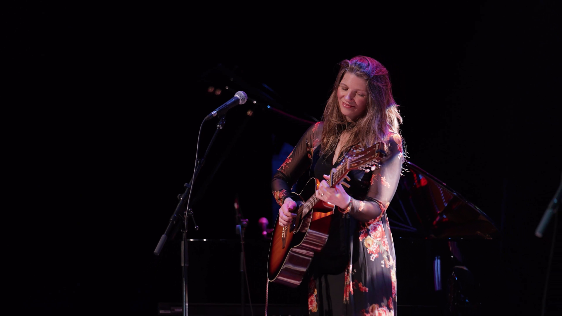 Musician in floral dress plays acoustic guitar on dark stage beside microphone stand.