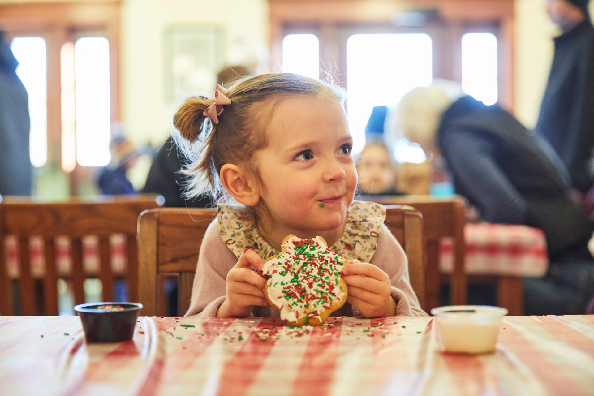 Child holds a large cookie decorated with colorful sprinkles at a holiday table.