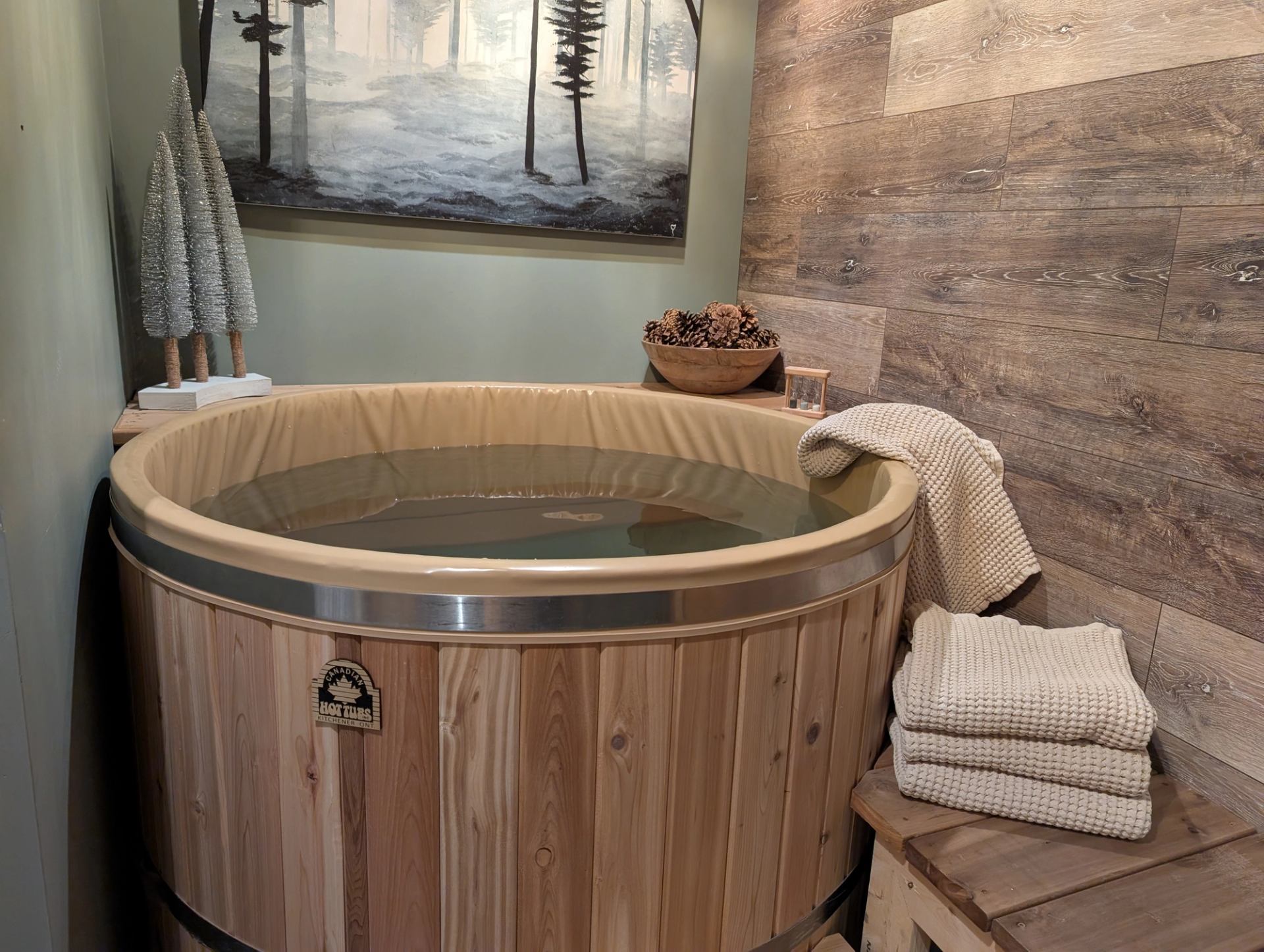 A classic wooden cold plunge tub filled with clear water sits tucked into a tranquil corner with wood-paneled walls. A neat stack of neutral-toned waffle towels rests on a bench next to the tub.