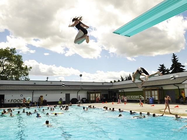 Swimmers enjoying a busy outdoor pool with diving boards and people jumping into the water.