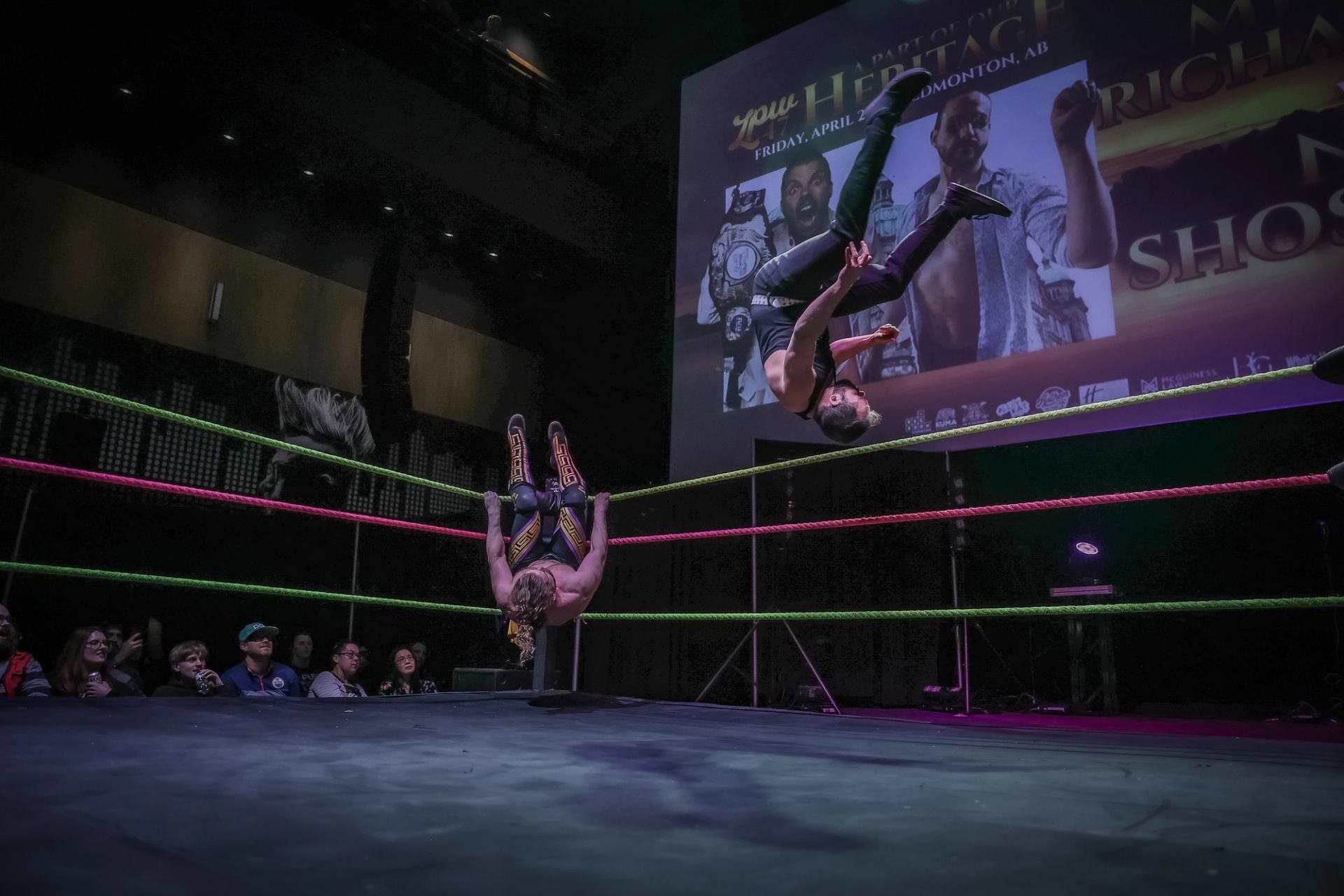Professional wrestlers flip in mid-air above ring ropes as audience watches and screen glows.