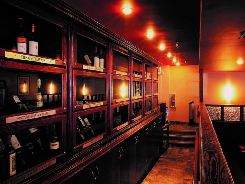 A warmly lit bar featuring a long wooden display cabinet filled with wine bottles and glowing lights.