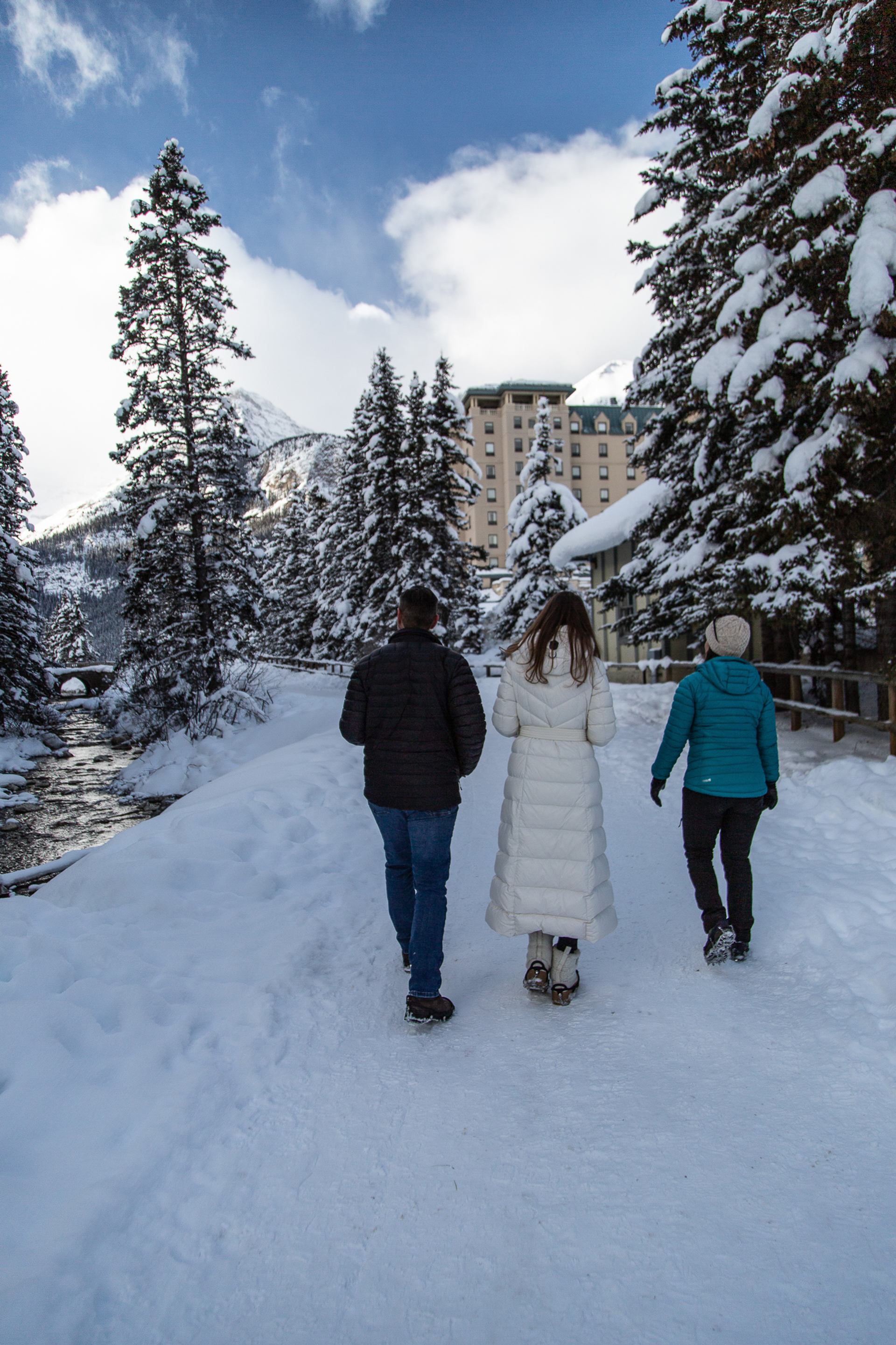 Three people walking on a snow-covered path lined with tall pine trees and a hotel in view