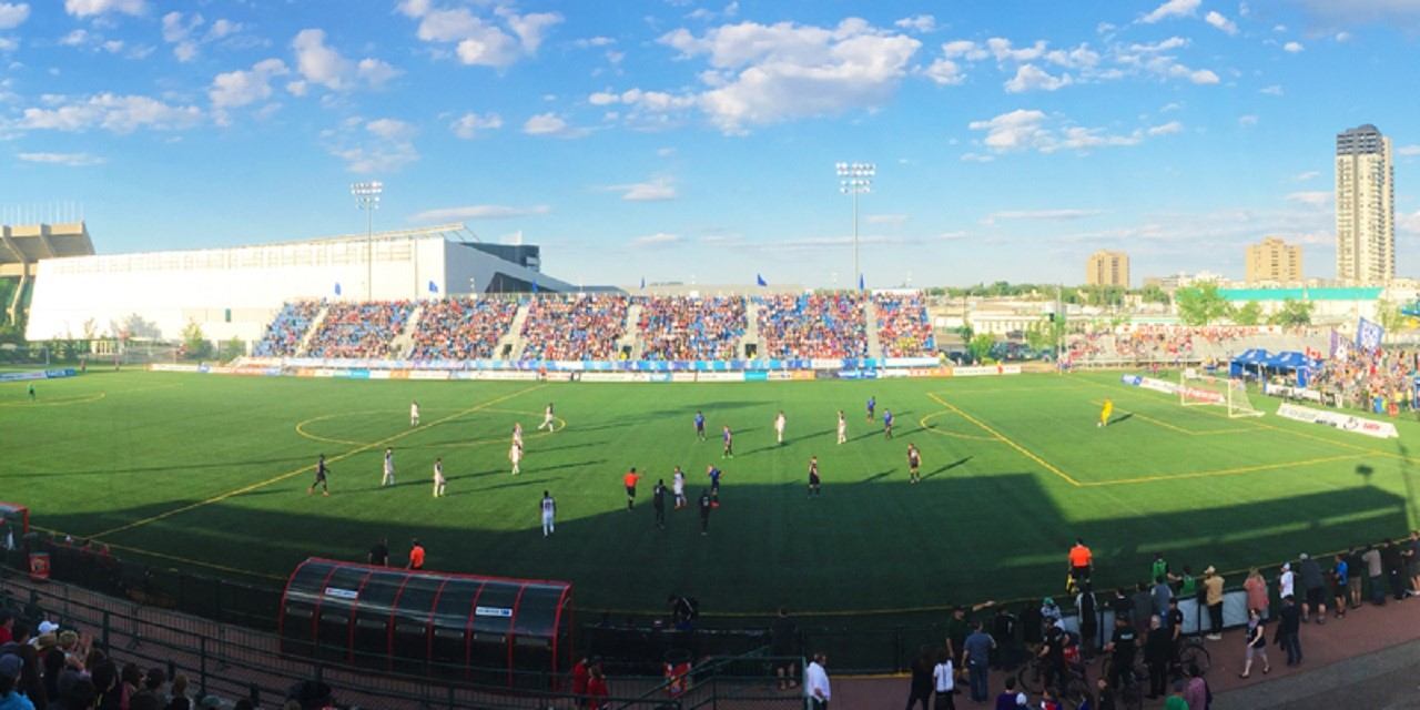 Soccer match at Clarke Stadium with players on the field and full stands under a bright blue sky.