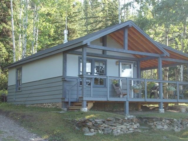 A grey cabin with a wide covered porch featuring a natural wood ceiling, surrounded by trees.