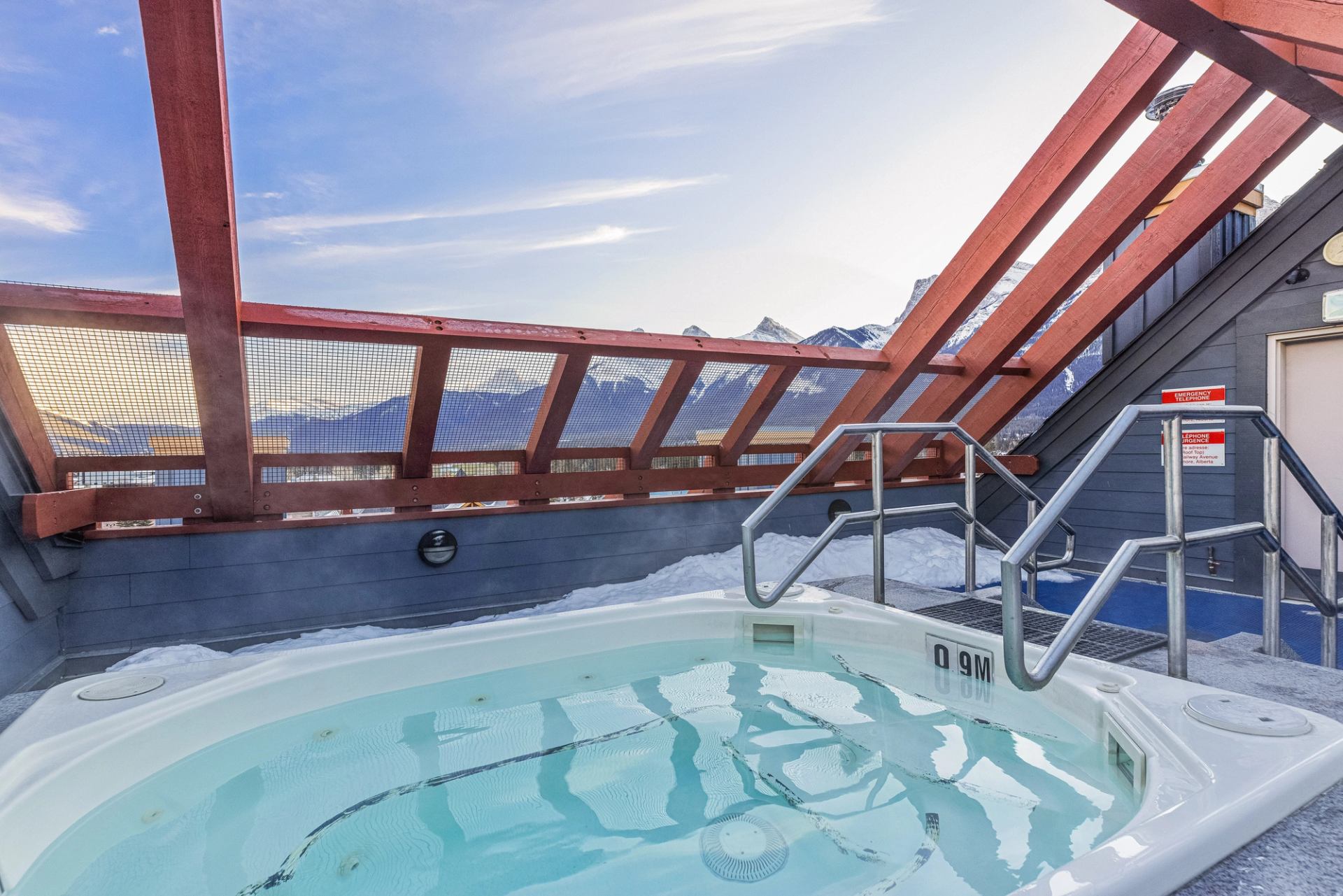 Rooftop hot tub with metal handrails, snow on deck, mountains visible through railing.