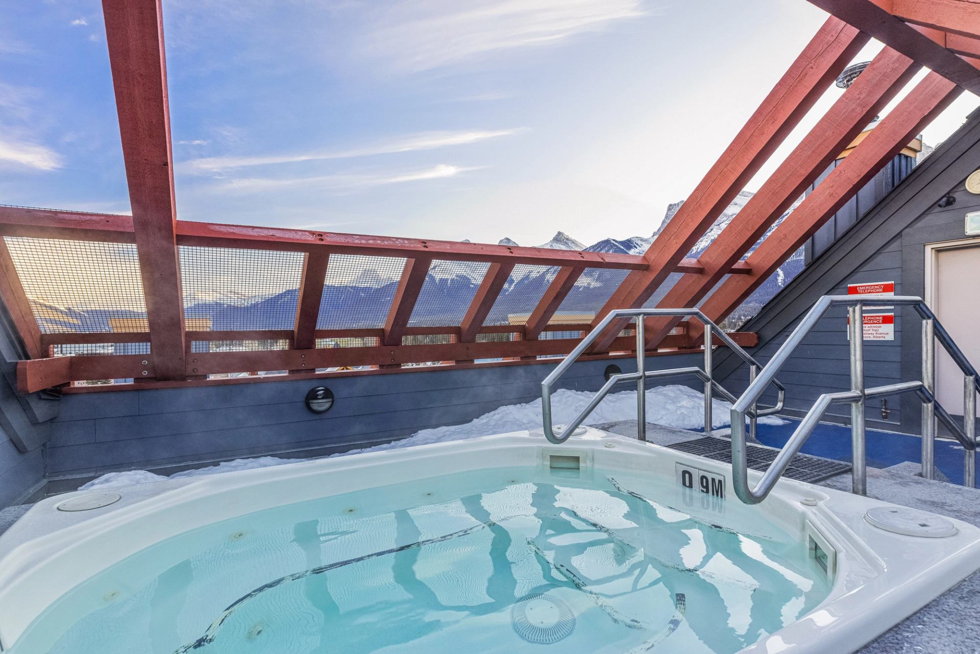 Rooftop hot tub with metal handrails, snow on deck, mountains visible through railing.
