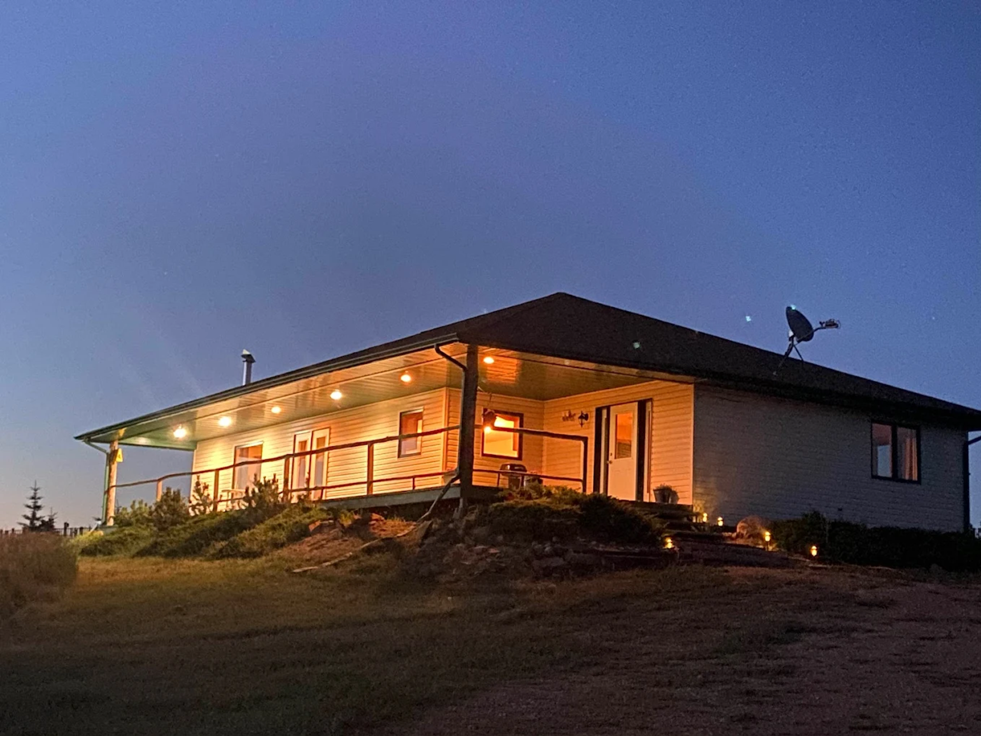 A warmly lit house at dusk with a wraparound porch and open sky above.