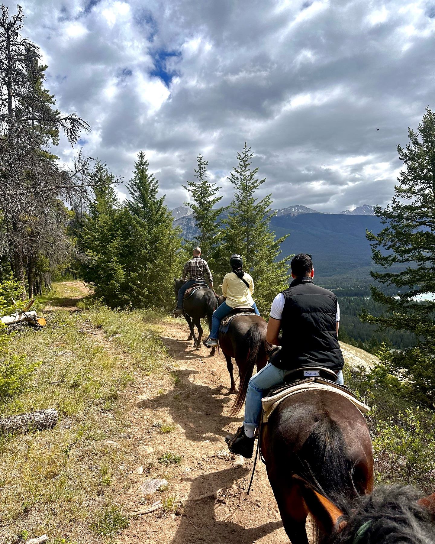 Horseback riders follow a dirt trail toward mountains under cloudy skies in Jasper.