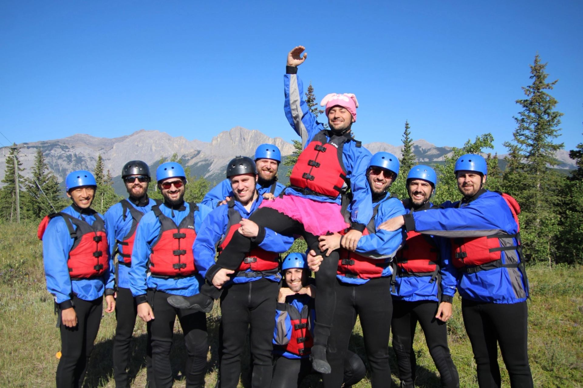 Group of rafters in helmets and lifejackets posing together in front of mountains.
